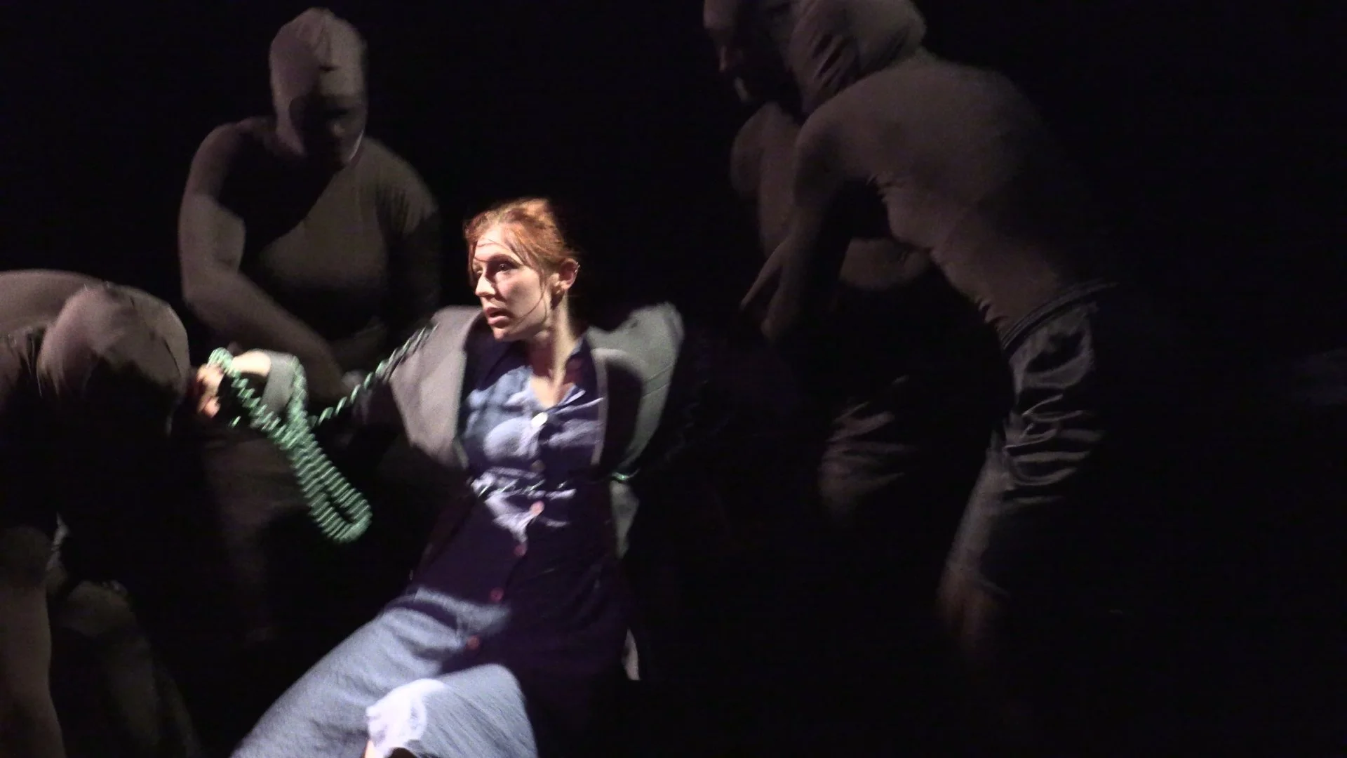 Woman in a dimly lit theatrical scene surrounded by masked performers and holding a rotary telephone.