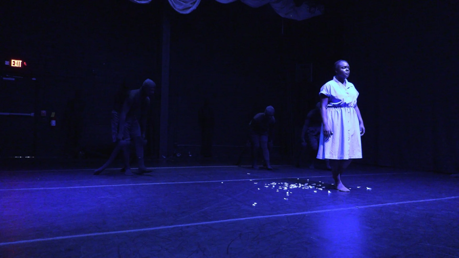 Blue-lit theatrical stage with a barefoot woman in a white dress, shadowy performers behind her, and scattered white debris on the floor.