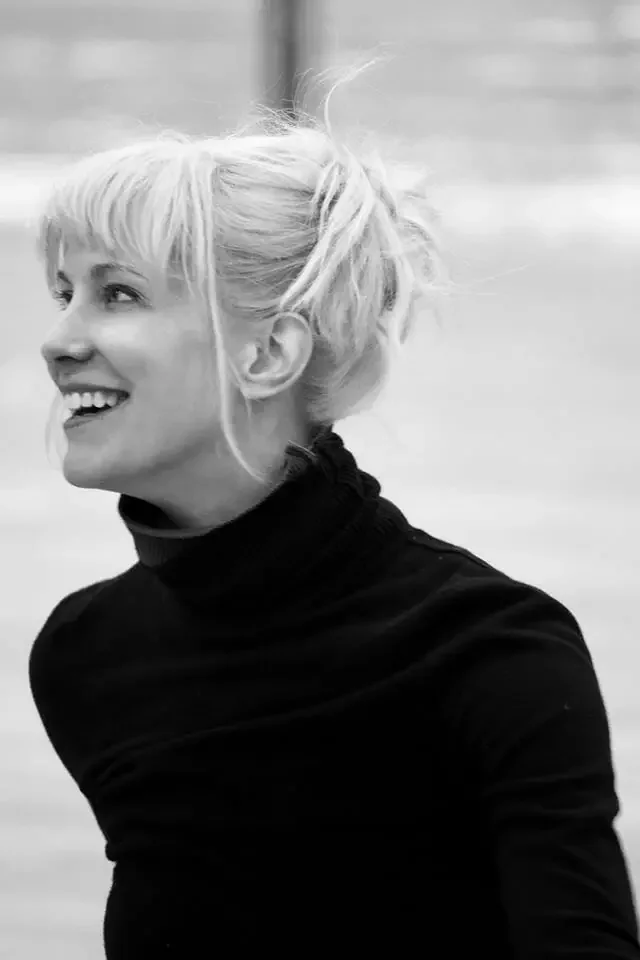 Black-and-white portrait of a smiling blonde woman with her hair in a messy bun, wearing a black turtleneck sweater.
