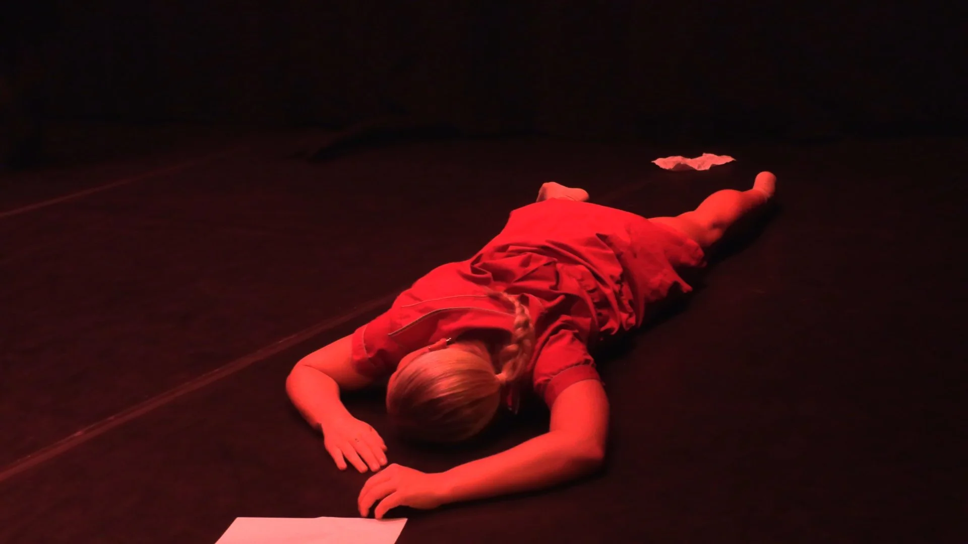 Dancer lying face down on a dark stage under red lighting.
