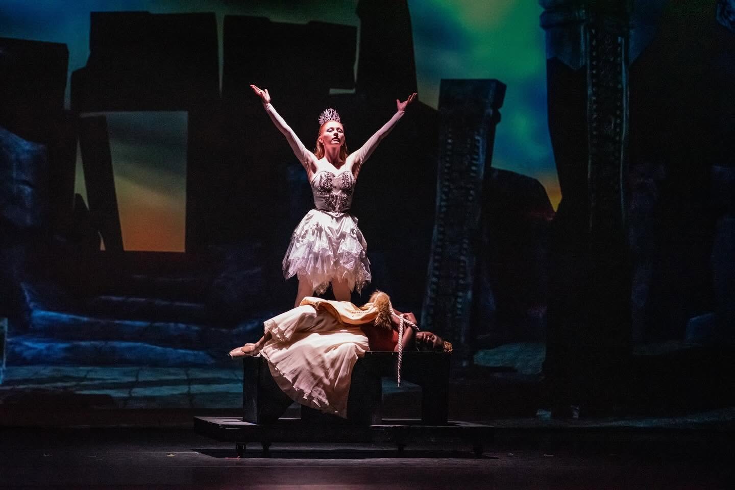 Theater performer in a crown and white costume raises her arms over a person lying across a bench on a dark stage.
