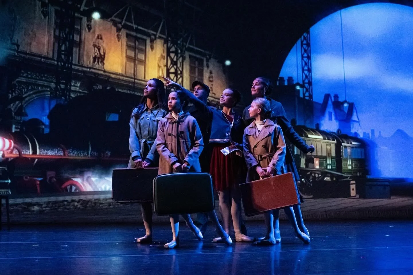 Young performers in vintage clothing hold suitcases on a theater stage with a blue-lit train station backdrop.