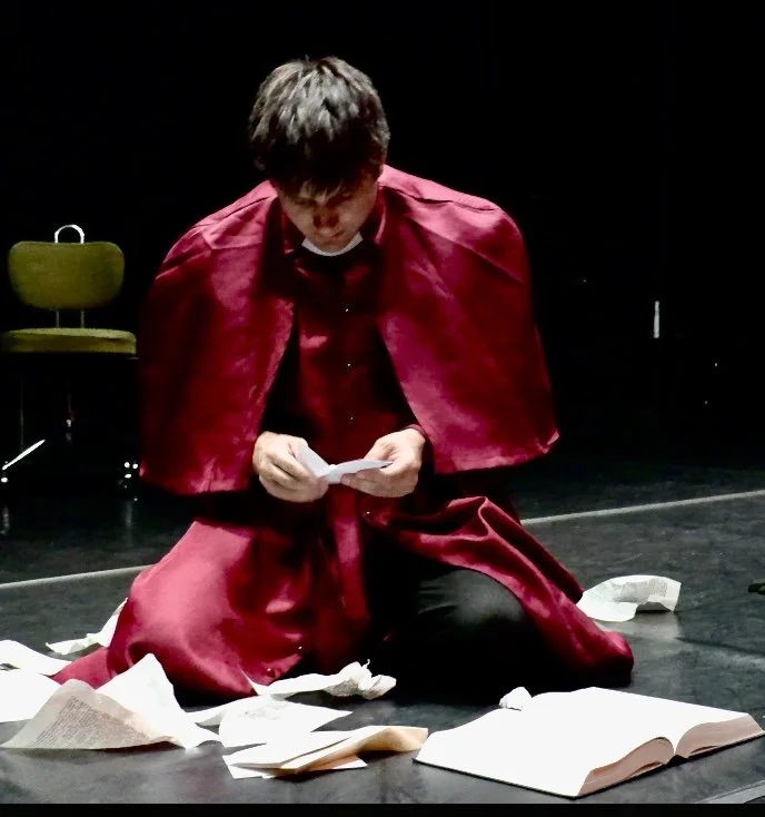 Actor in a red cape kneeling on a stage and reading a paper, surrounded by scattered books and papers.