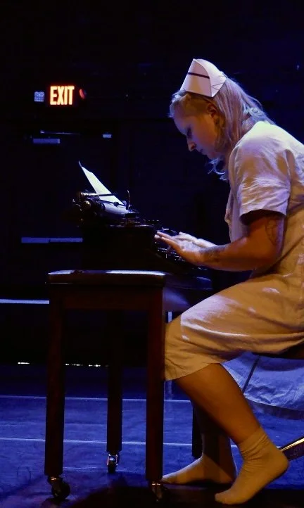 Performer in a white costume and nurse hat uses a typewriter on stage under blue lighting.