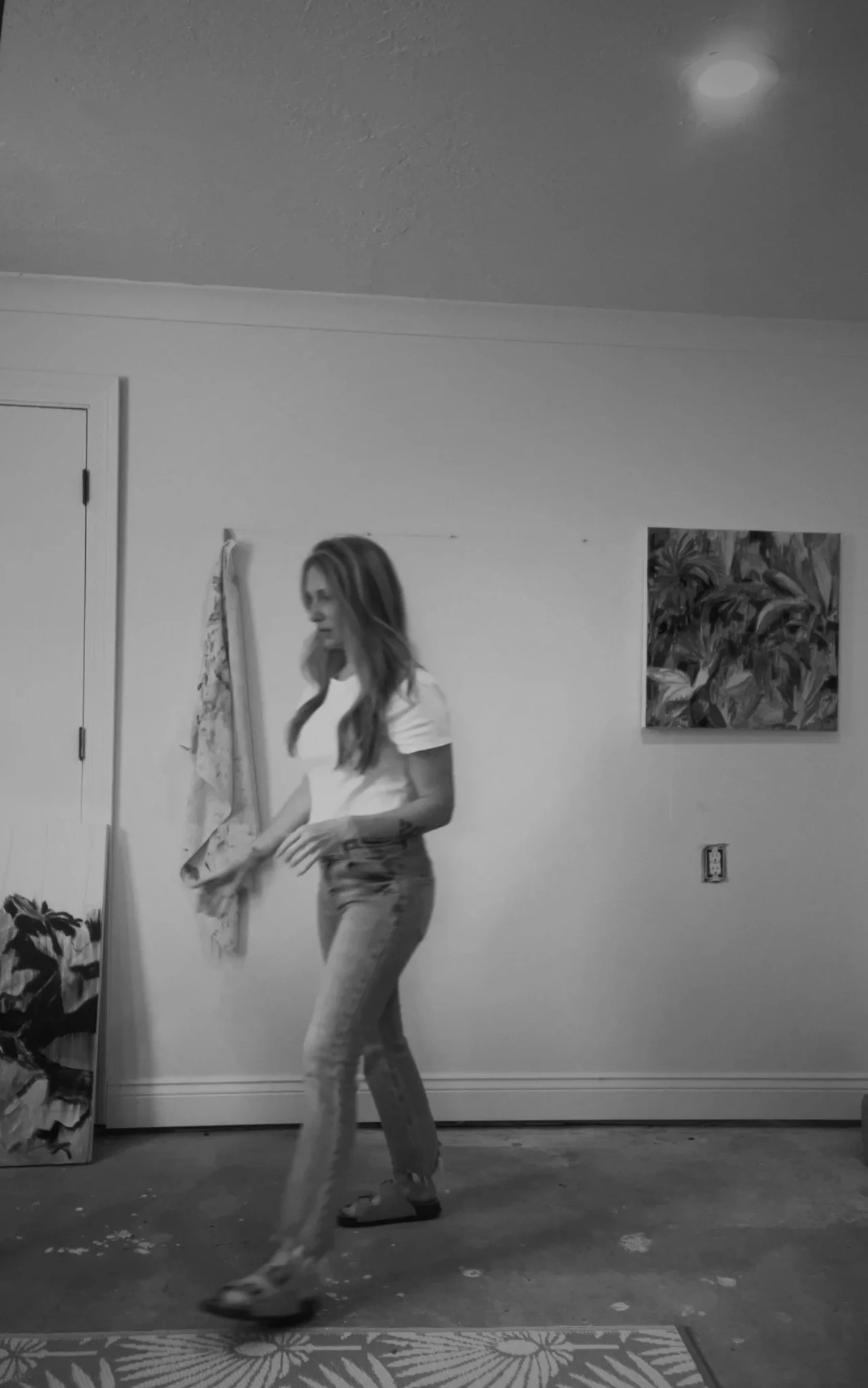 A woman with long hair walking barefoot on a mat inside a room with a white wall, a door, artwork, and a plant.