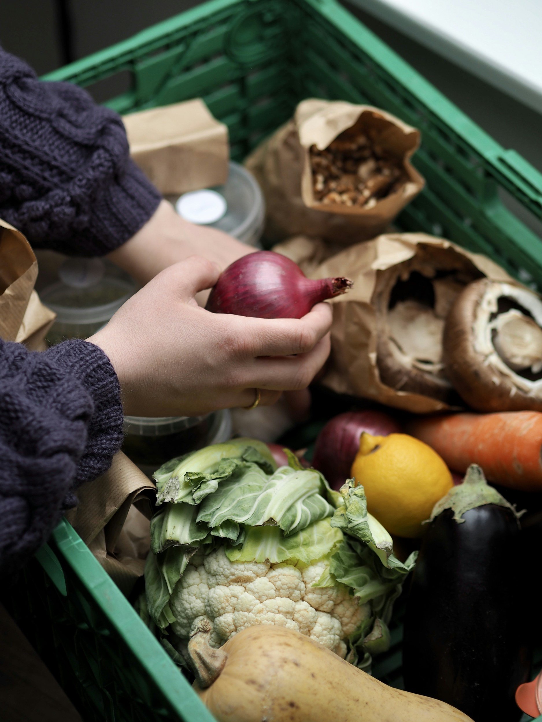 A green shopping basket filled with fresh vegetables including cauliflower, a red onion, a yellow lemon, a large orange carrot, a butternut squash, and green lettuce.