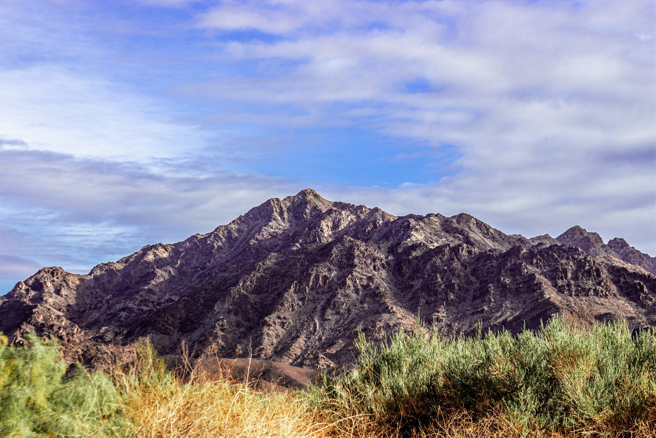 An Arizona mountain range with rugged peaks under a partly cloudy sky, with desert vegetation in the foreground.