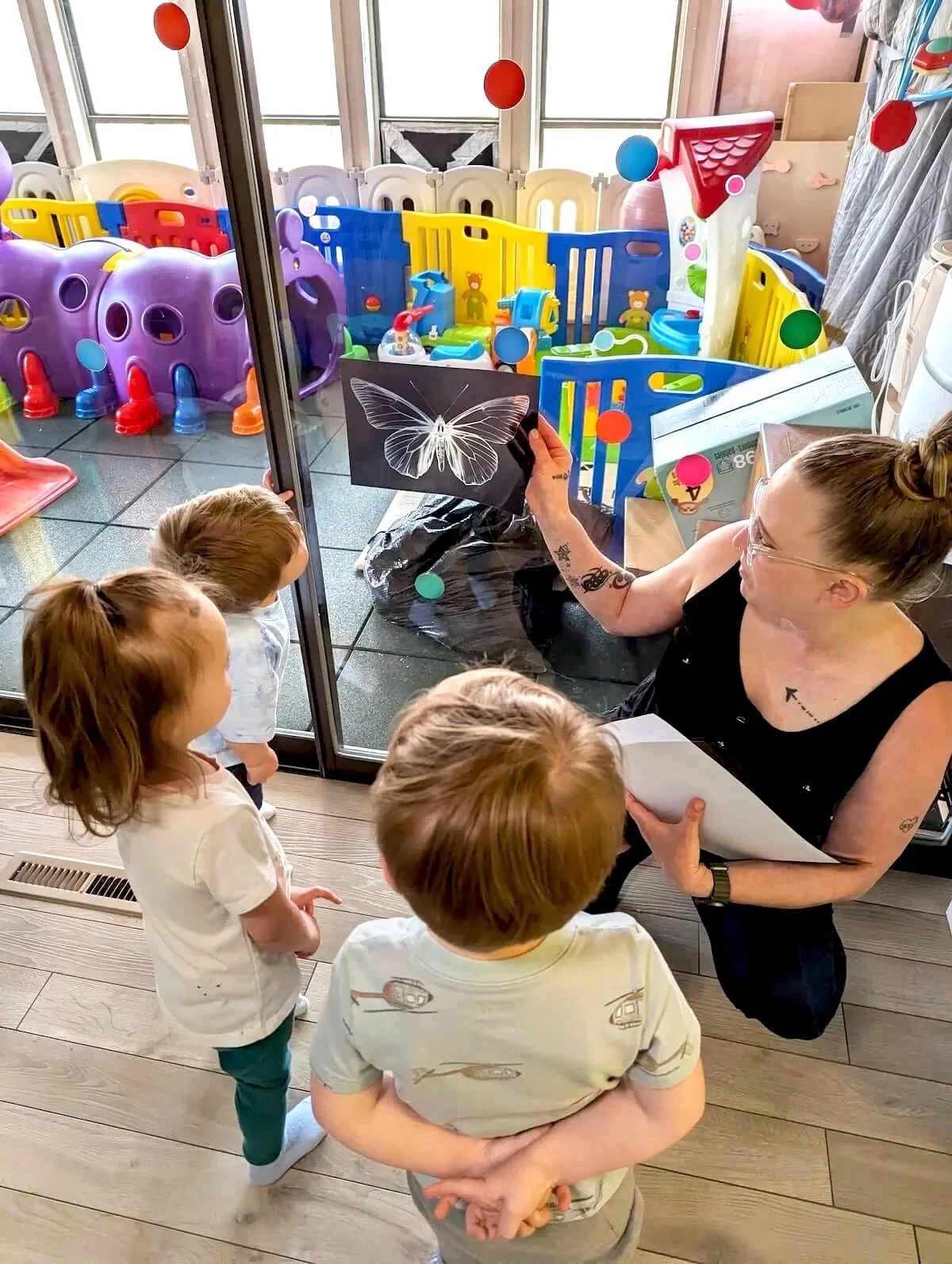 A woman teaching a group of young children about a butterfly illustration inside a room with a glass door. The children are gathered around her, with the room visible through the door, showing colorful play equipment in a fenced area.