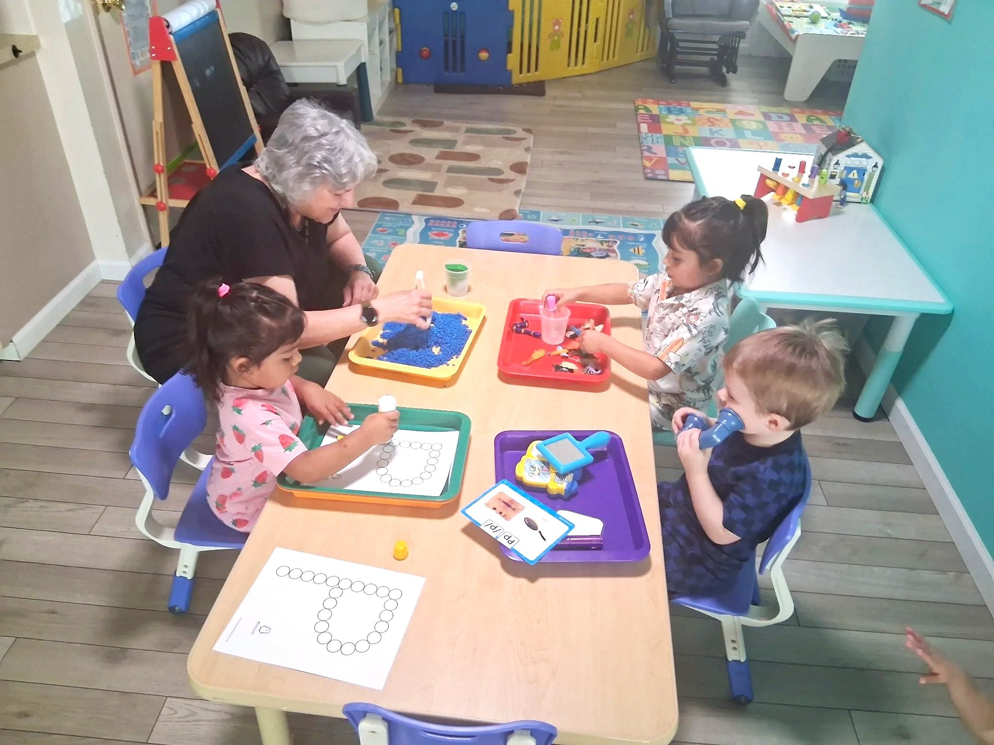 A classroom with an adult and five children sitting around a table and engaging in activities such as coloring, playing with blue kinetic sand, and using clips. The children are working on papers with circles, and various educational items like a toy house and alphabet cards are visible in the room.