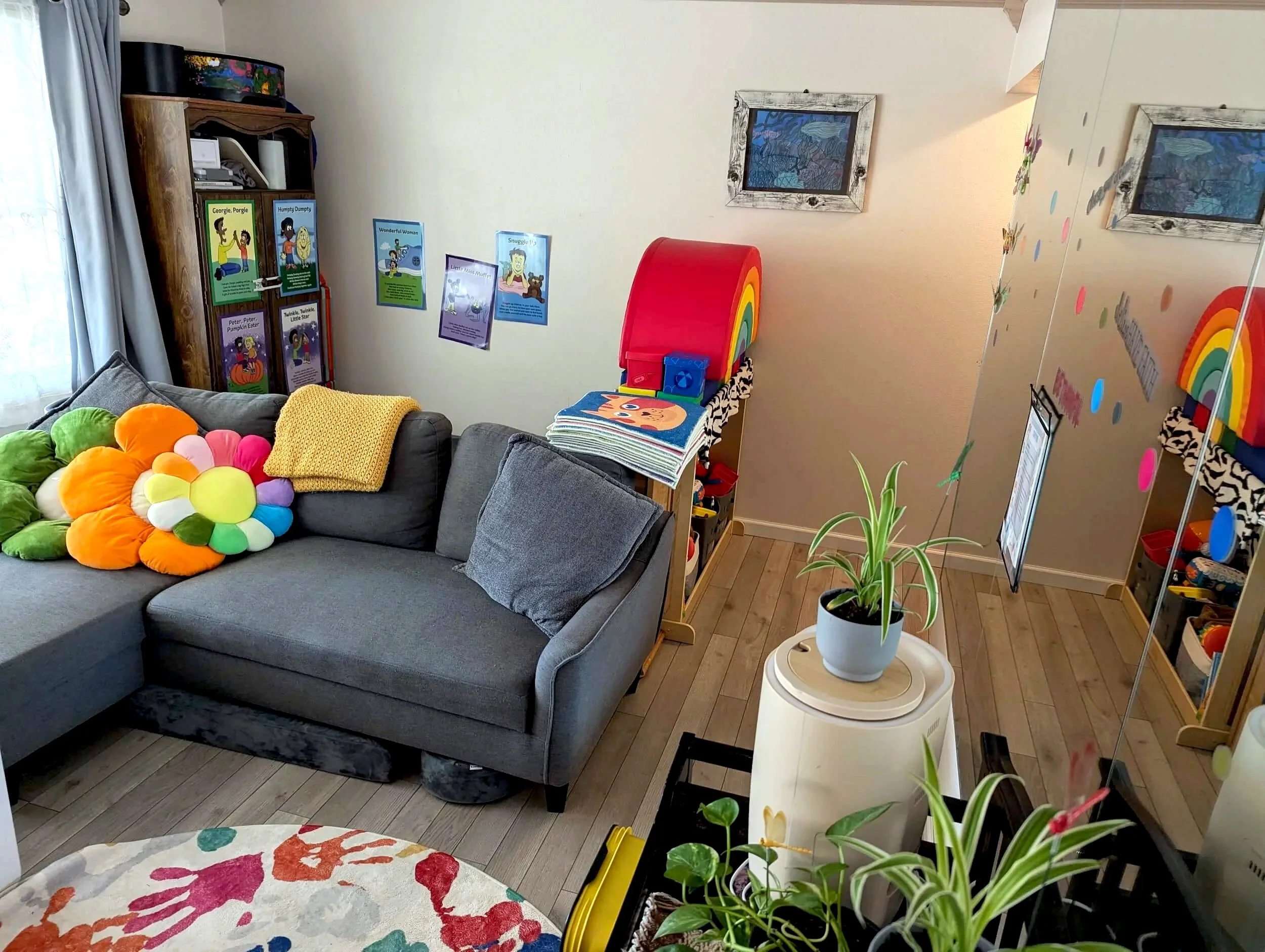 A room decorated for children with a gray sofa, colorful plush flower pillow, bookshelf with children's books, and a colorful rainbow storage container. Potted plants and children's artwork are also visible.