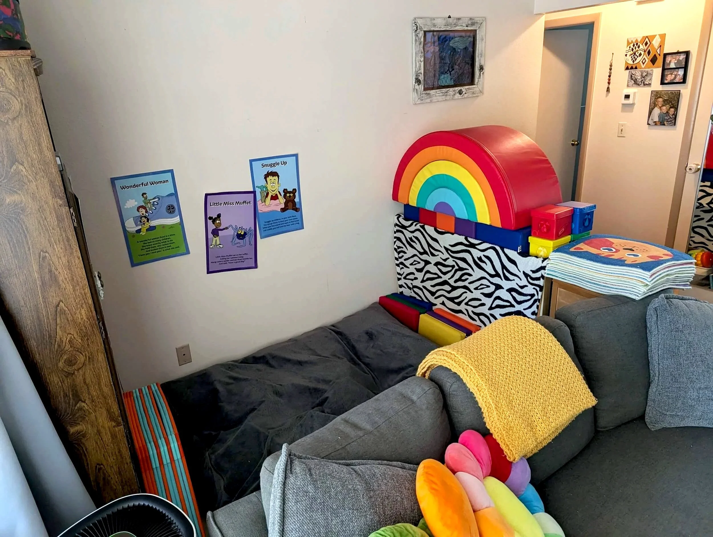 A cozy living room with a gray couch covered with a black blanket and colorful plush toys on it. A yellow knitted blanket is draped over the couch. There is a wooden side table and a zebra-print tablecloth with rainbow-shaped foam padding arranged on top. The wall has framed pictures and posters, including a set of illustrated story posters with titles 'Wonderful Woman,' 'Little Miss Muffet,' and 'Snuggle Up.' A colorful striped mat is on the floor, and a rainbow-shaped foam play structure is against the wall.
