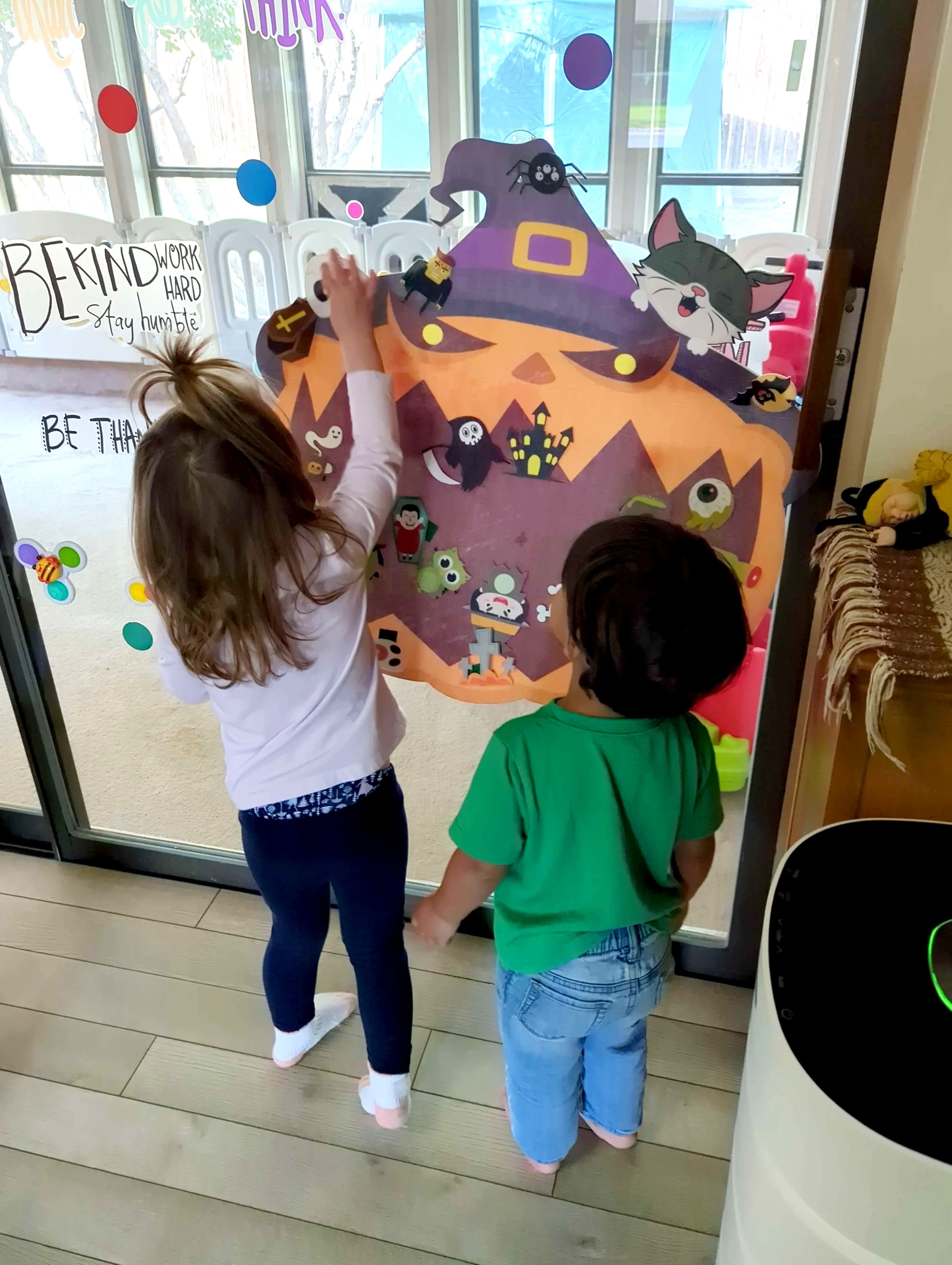 Two children standing at a glass door with Halloween-themed decorations, including a large orange pumpkin cutout with a witch's hat and various small Halloween stickers, as they appear to be knocking or looking inside.