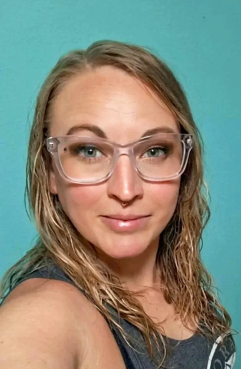 A woman with wavy blonde hair, wearing clear glasses, looking at the camera with a slight smile, against a teal background.