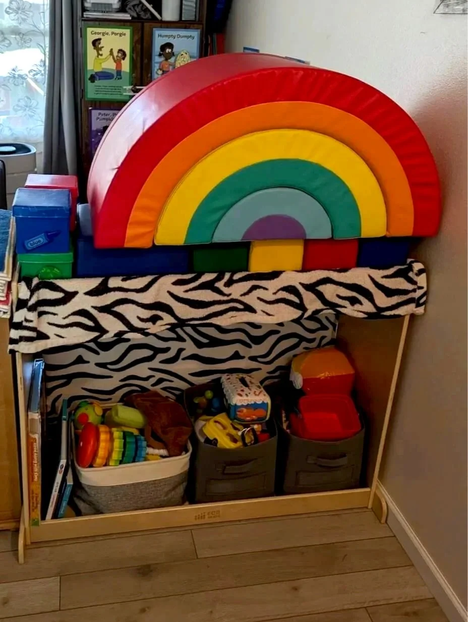 A rainbow-shaped wooden toy with red, orange, yellow, green, blue, and purple arcs. The rainbow is placed on a shelf with zebra-print fabric. Below the shelf are various toys and books in bins. Behind the rainbow, there are children's books and a curtain.