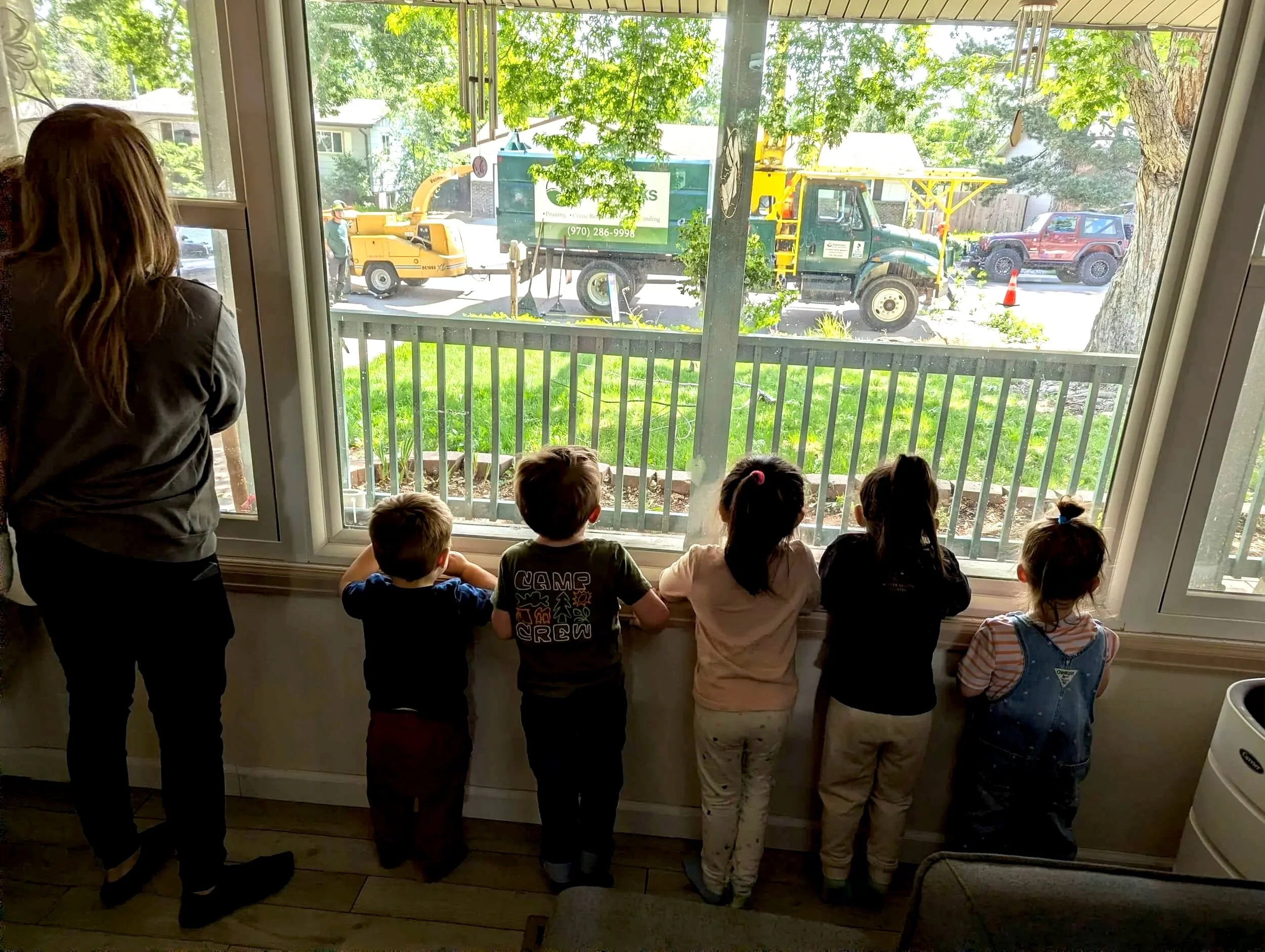 Children watching a tree-trimming truck outside through large window in a room with wooden flooring and a woman with long hair standing on the left.