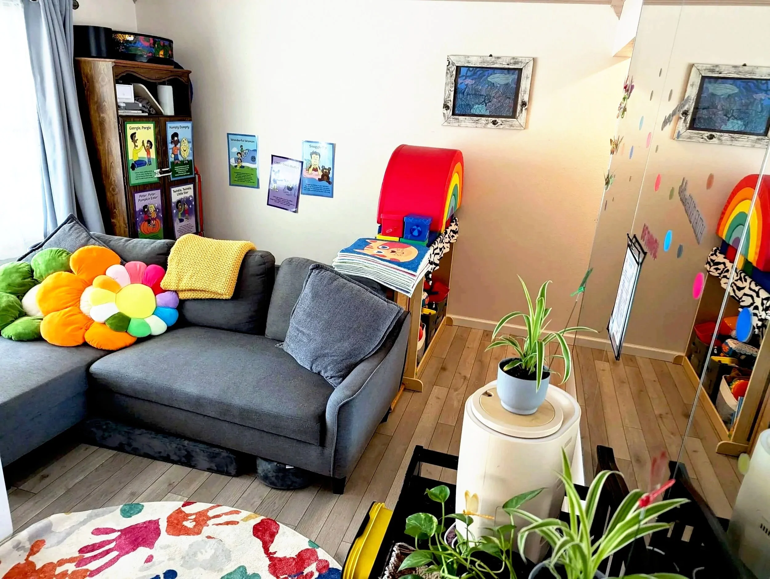 Living room with gray sectional sofa decorated with colorful plush flower pillows, a white side table with a green potted plant, a wooden entertainment center displaying children's books, colorful posters on the wall, and a corner with rainbow-themed decorations and plants.