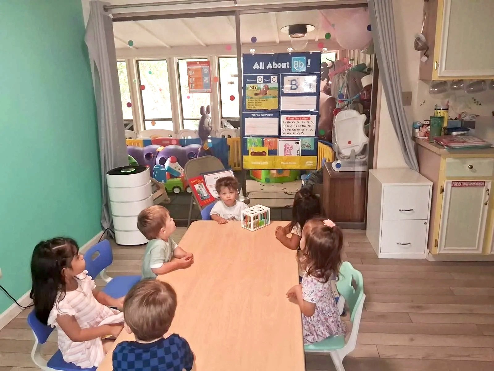 Children sitting around a table in a classroom or daycare setting, with colorful decorations and educational posters on the walls, and outdoor toys visible through the windows in the background.