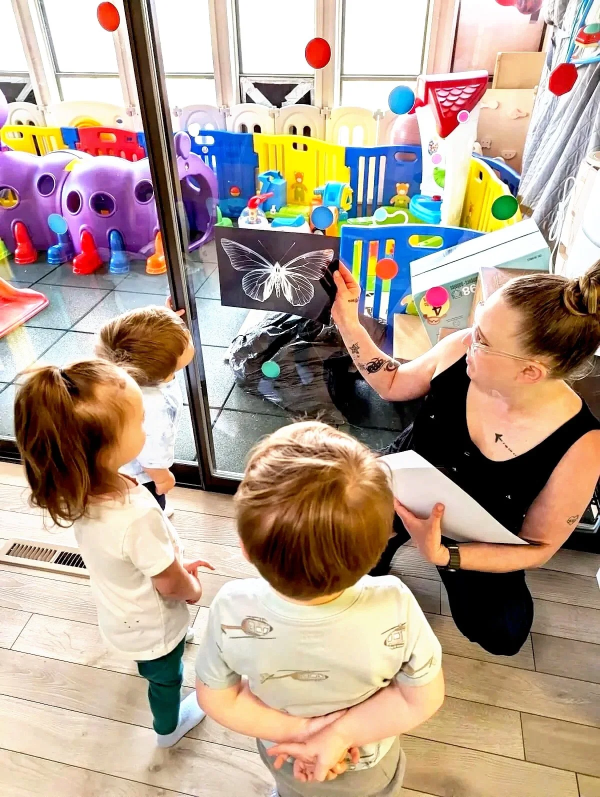 A woman showing a butterfly drawing to three children inside a house, with a view of a colorful outdoor play area with plastic fences and toys through a glass door.