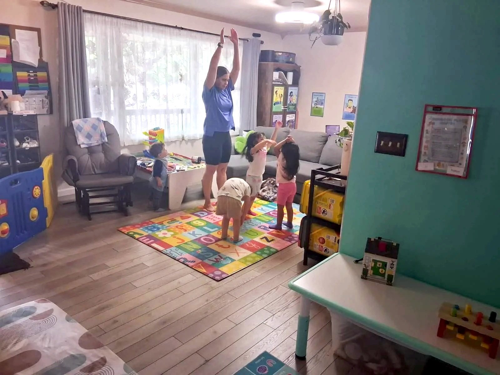 An indoor scene with a woman and five children on an alphabet and number rug, engaging in a stretching or dance activity. The room has a couch, a chair, a window with curtains, and various children's toys and books, suggesting it's a playroom or classroom setting.