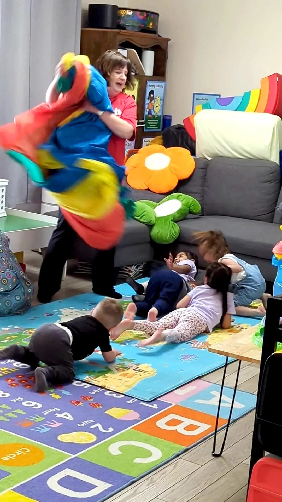 A woman is playing with a colorful parachute inside a room filled with children on a playmat. Some children are lying on their stomachs, and the room has large plush flowers on the sofa and educational posters on the wall.