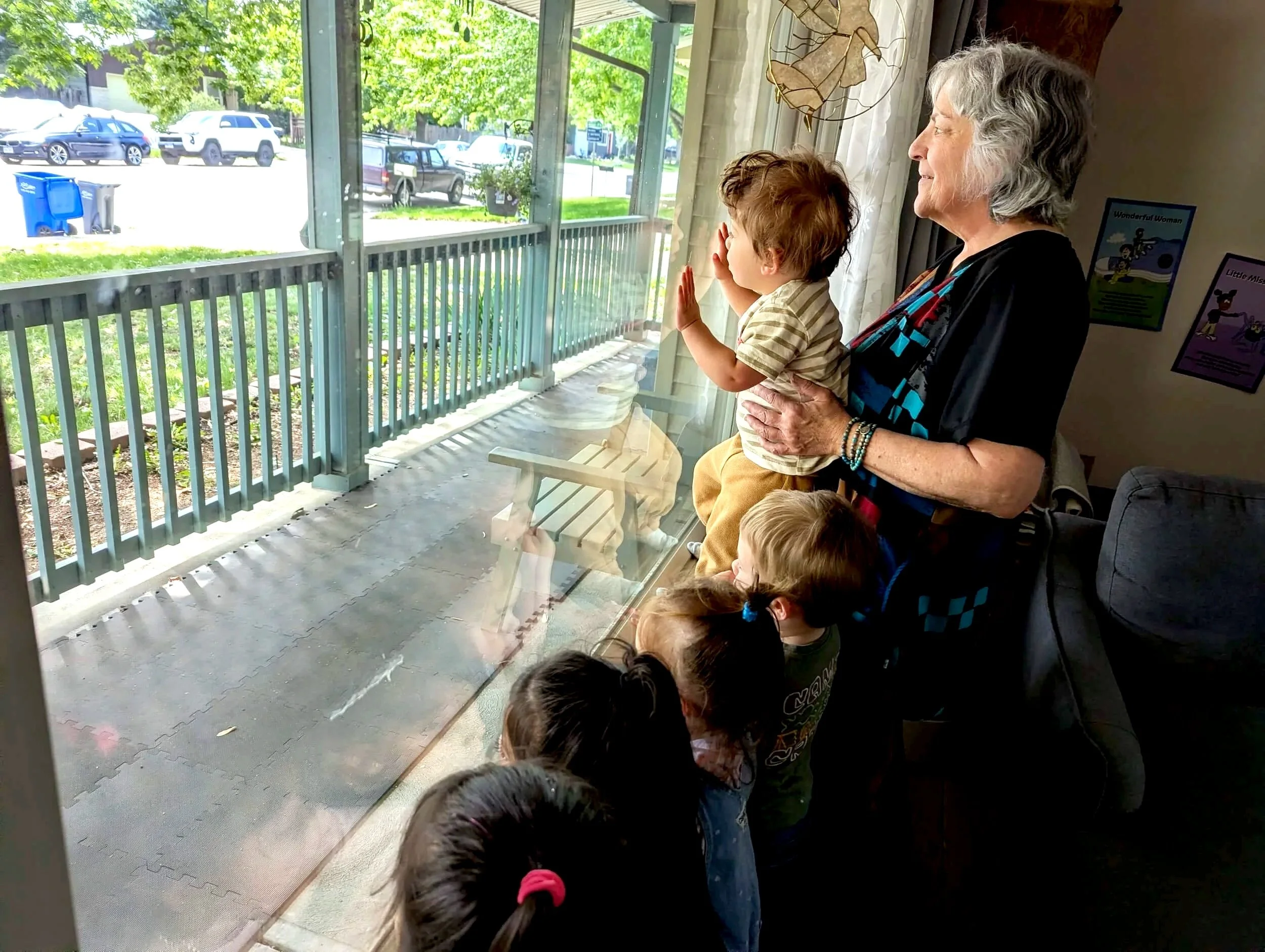 A group of children and an older woman are looking through a window at something outside. The children are standing close to the window, and the woman is holding one of the children. The scene appears to be inside a house or a classroom, with posters on the wall and a view of parked cars and greenery outside.