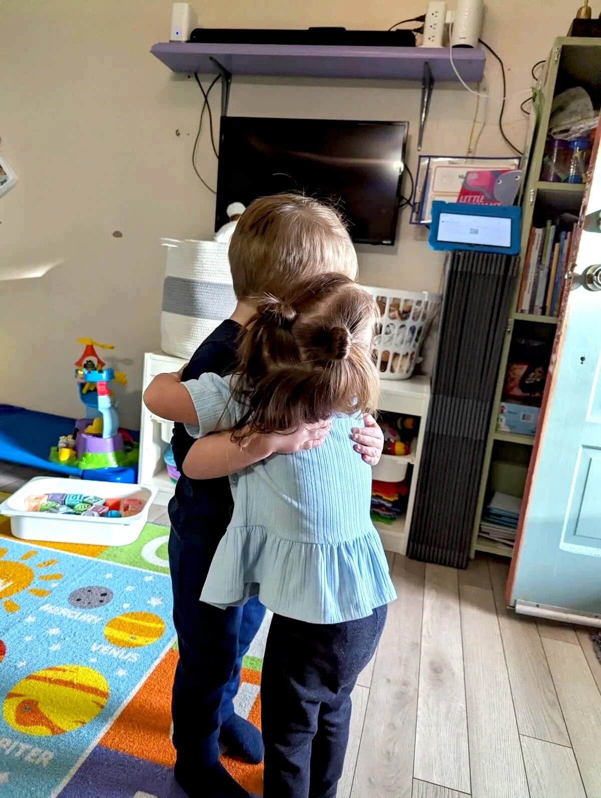 Two young children hugging each other in a living room with colorful toys, books, a television, and a patterned carpet.