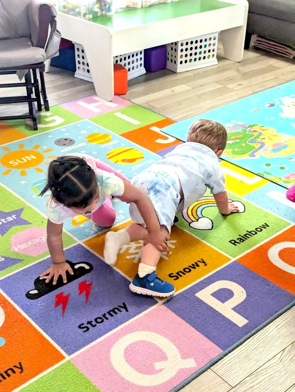 Two young children crawling on a colorful educational rug with animal and weather illustrations.
