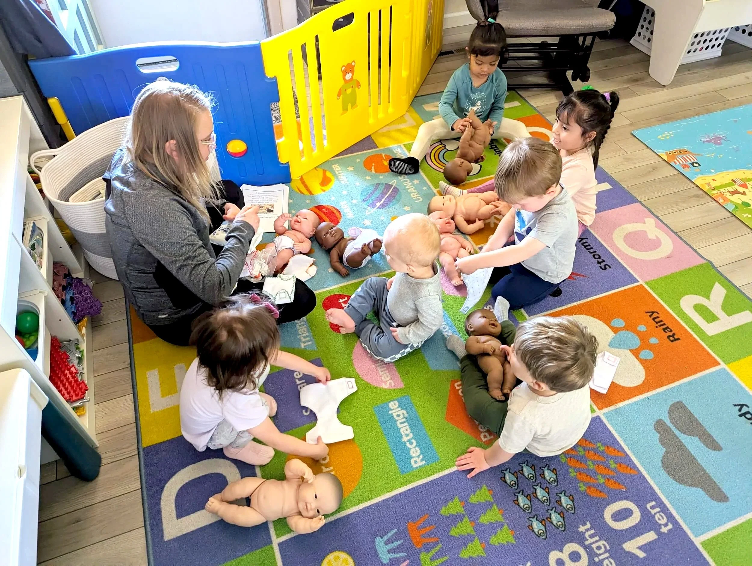 Children and infants sitting on a colorful educational carpet, engaging in an activity involving stuffed baby dolls, with a woman reading to them. The room has a bright, cheerful atmosphere with toys and furniture suitable for young children.