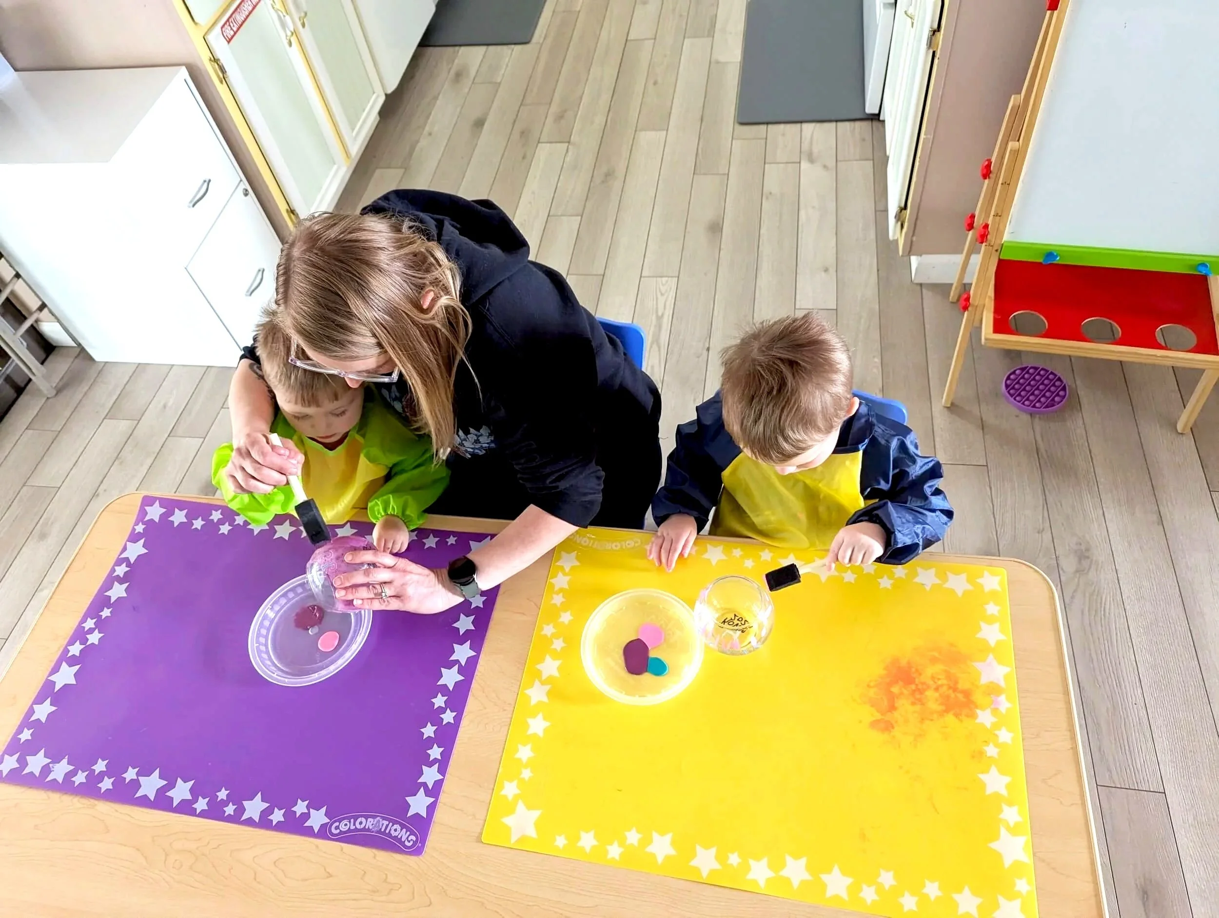 Two children and an adult sitting at a table engaging in a coloring activity. The table has colorful mats with star patterns, and there are small containers with colored foam shapes. In the background, a white cabinet, a gray mat, and a white stove are visible.