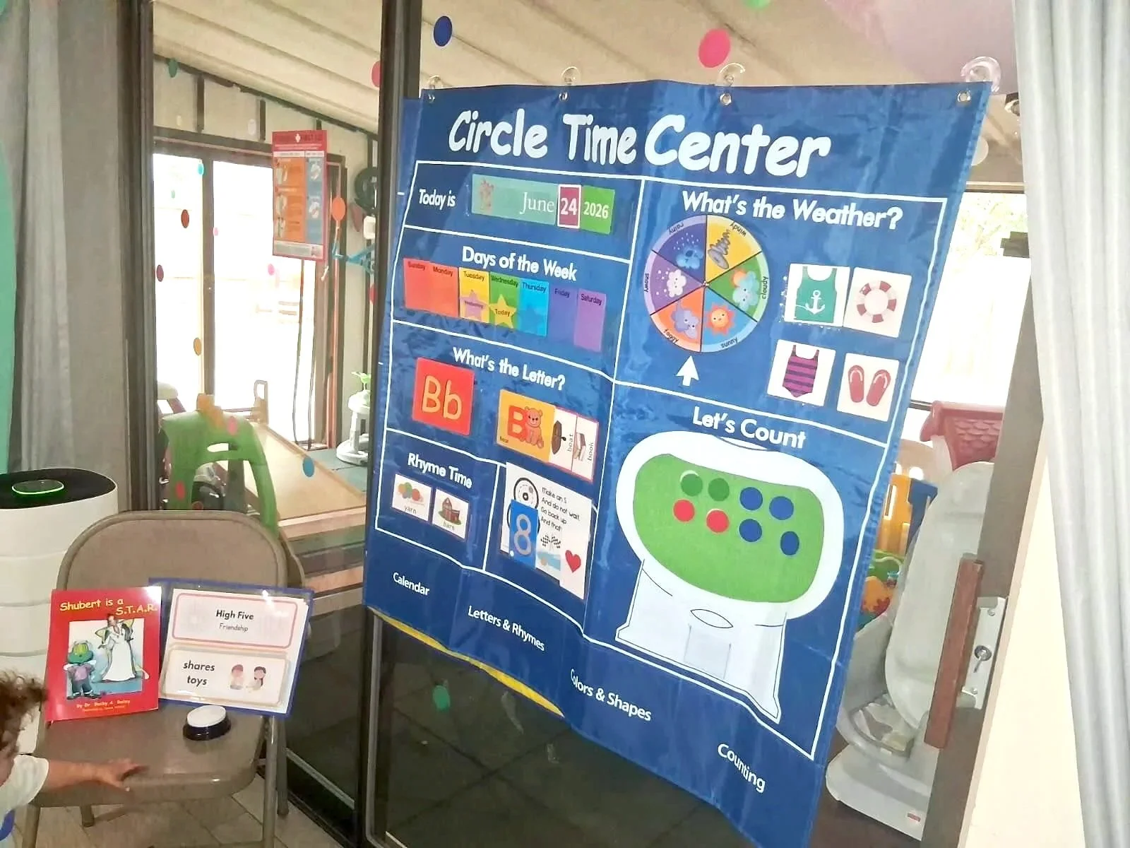 Educational poster titled 'Circle Time Center' hanging inside a classroom. The poster displays the date June 24, 2026, and includes sections about the weather, days of the week, alphabet letters, and counting. There are also some books and toys in the background.