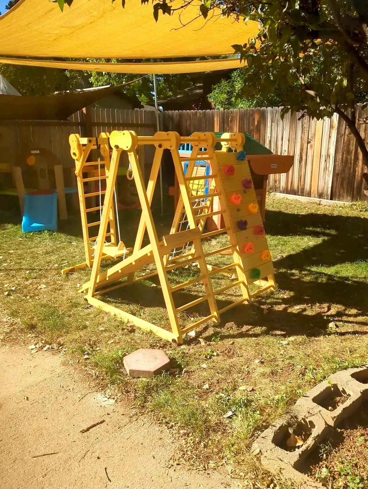 Child's backyard playground with yellow climbing gym, swings, and slide, shaded by a large yellow fabric canopy, surrounded by a wooden fence and trees