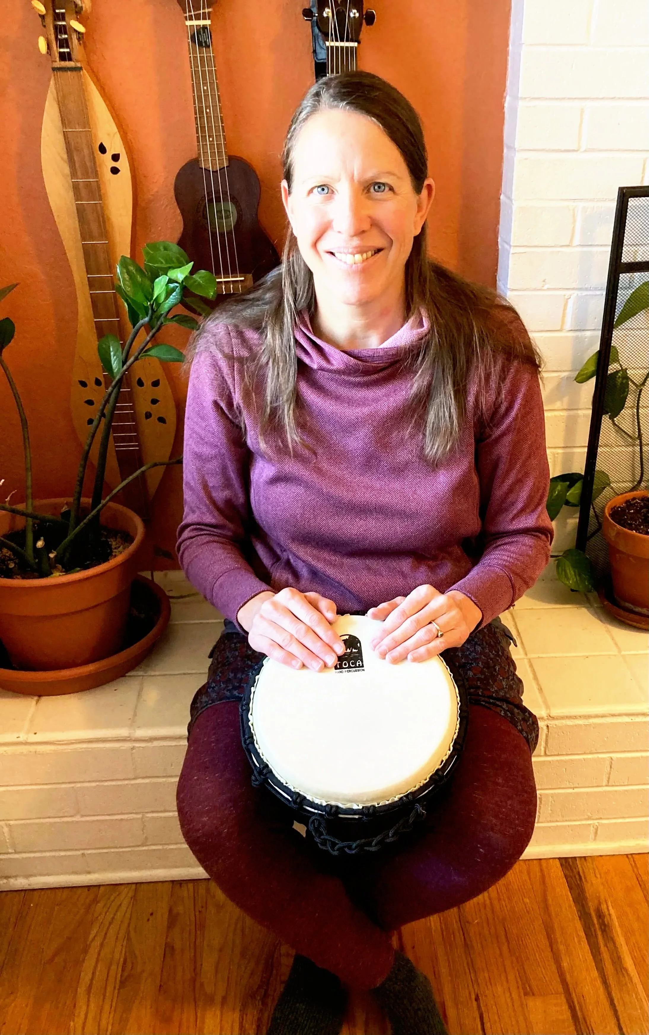 A woman sitting cross-legged on a wooden floor, holding a bong with a drumhead logo, smiling at the camera. Behind her, there are several ukuleles hanging on a wall and a potted plant next to her.