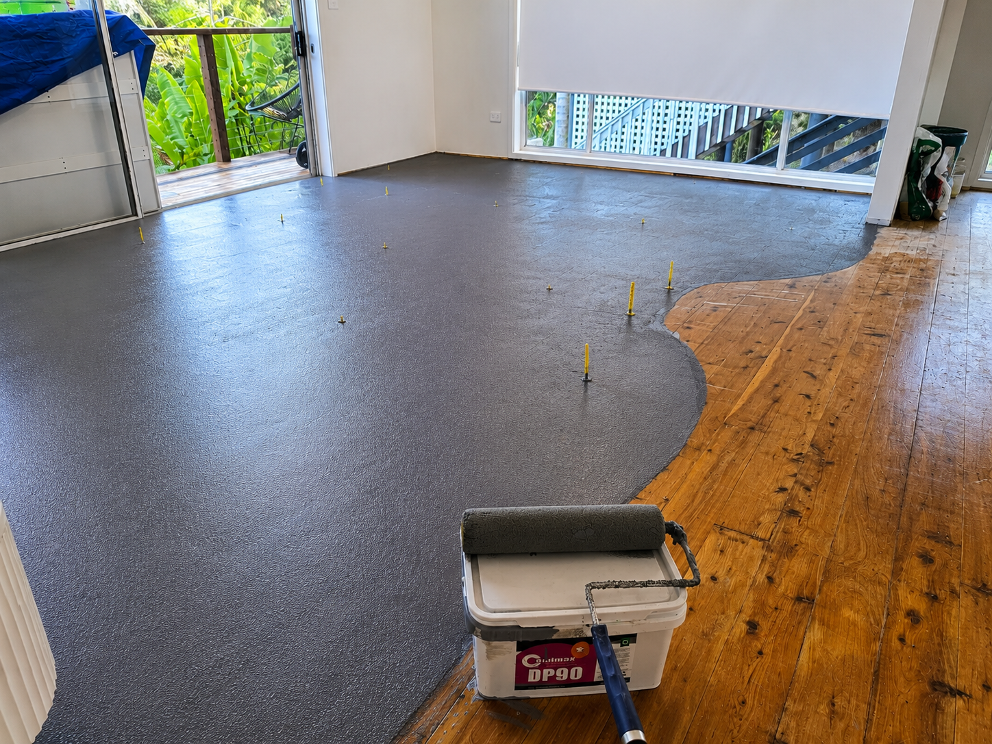 A room with a partially installed gray epoxy floor coating, with yellow markers indicating the areas of work. There is a bucket with a roller in the foreground, and a sliding door open to a balcony with green foliage outside.