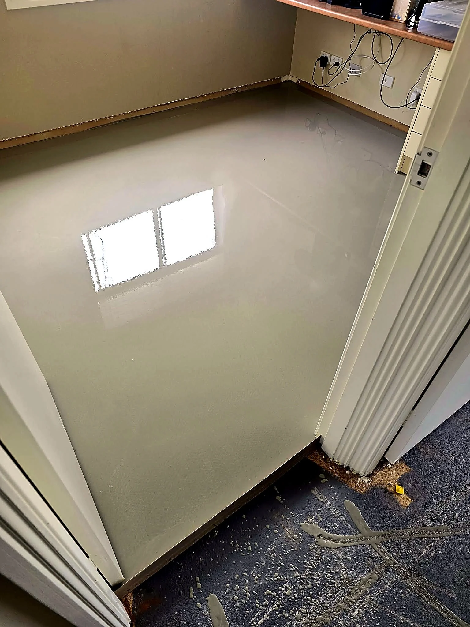 A room with a clean shiny floor, a wall with electrical outlets and cords, a brown wooden ledge with office supplies, and a door frame with chipped paint and debris on the floor.