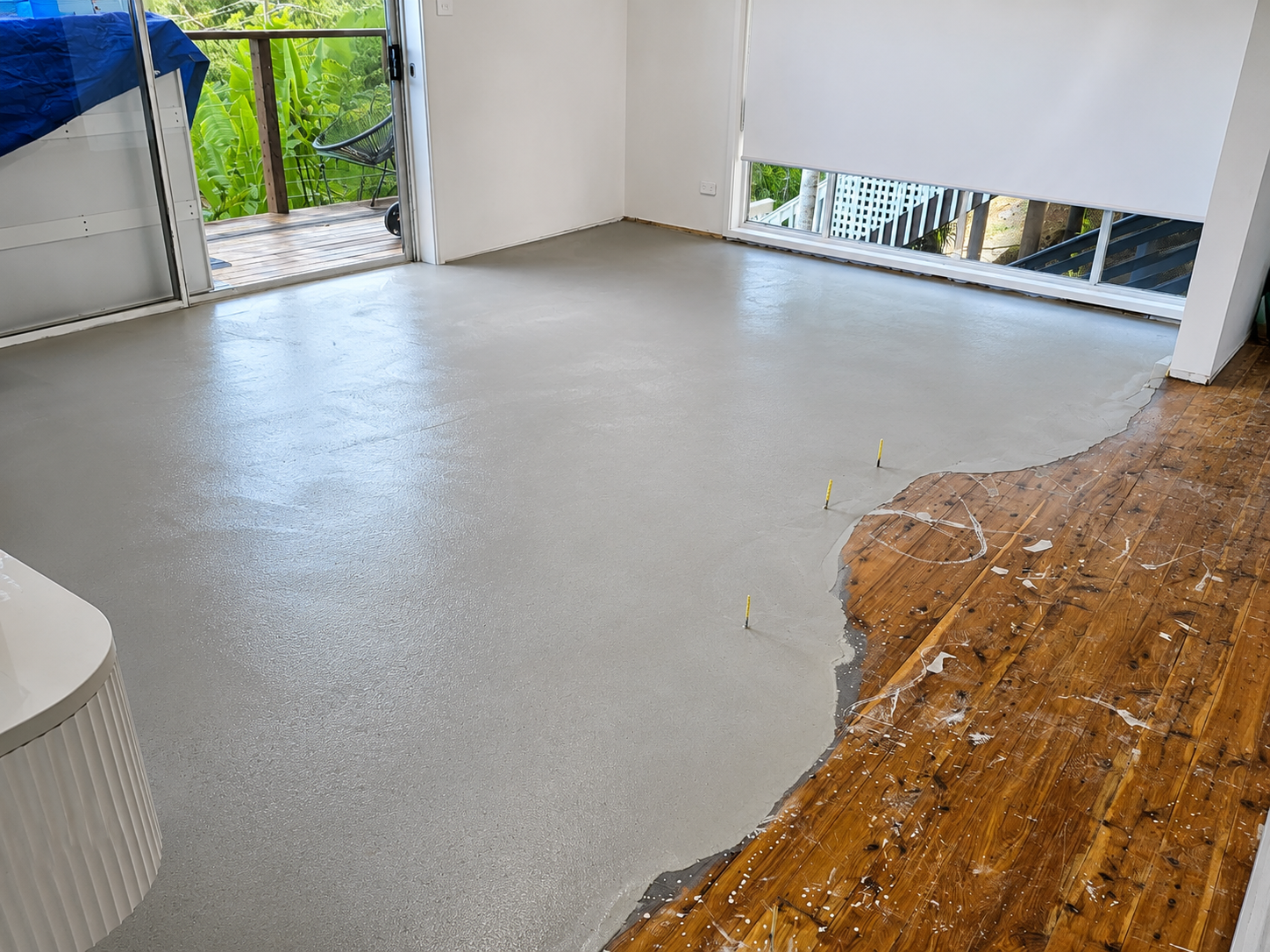 Living room with fresh concrete floor being poured over old wooden flooring, with patio and greenery visible through a partially open door.