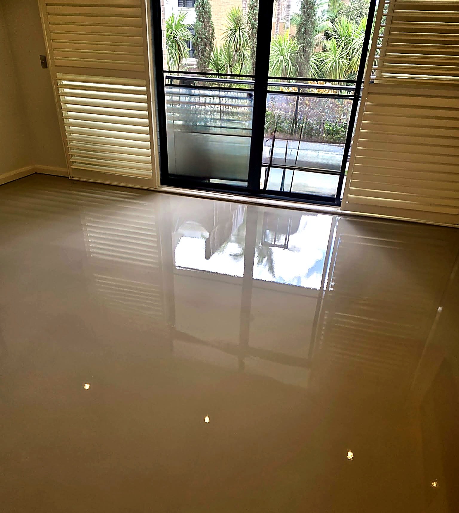 Living room with light-colored polished floor reflecting sunlight from sliding glass door, which opens to a lush outdoor garden with trees and bushes.