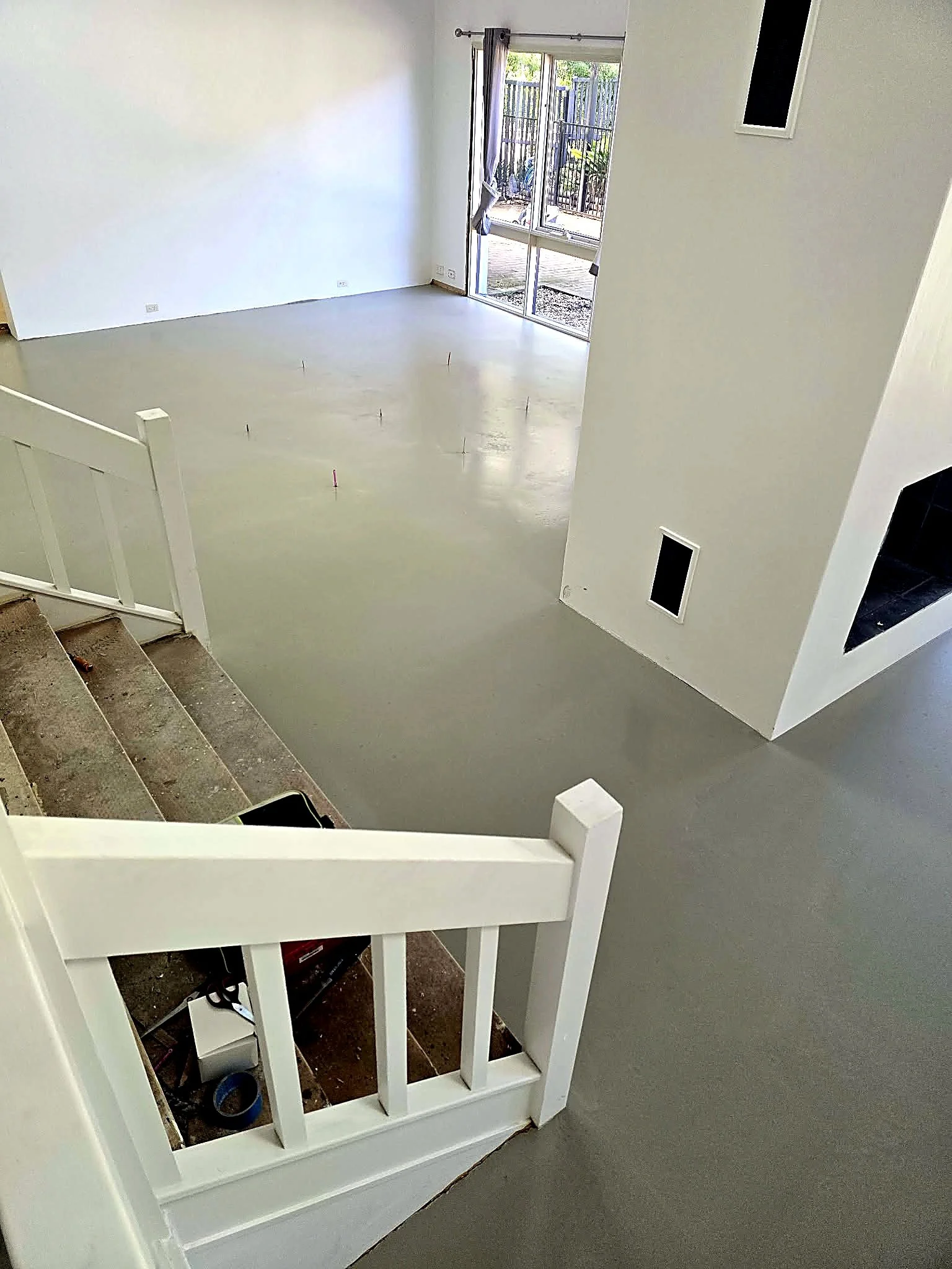 Empty living room with large window, sliding door, and staircase railing, and construction tools visible in the walking space.