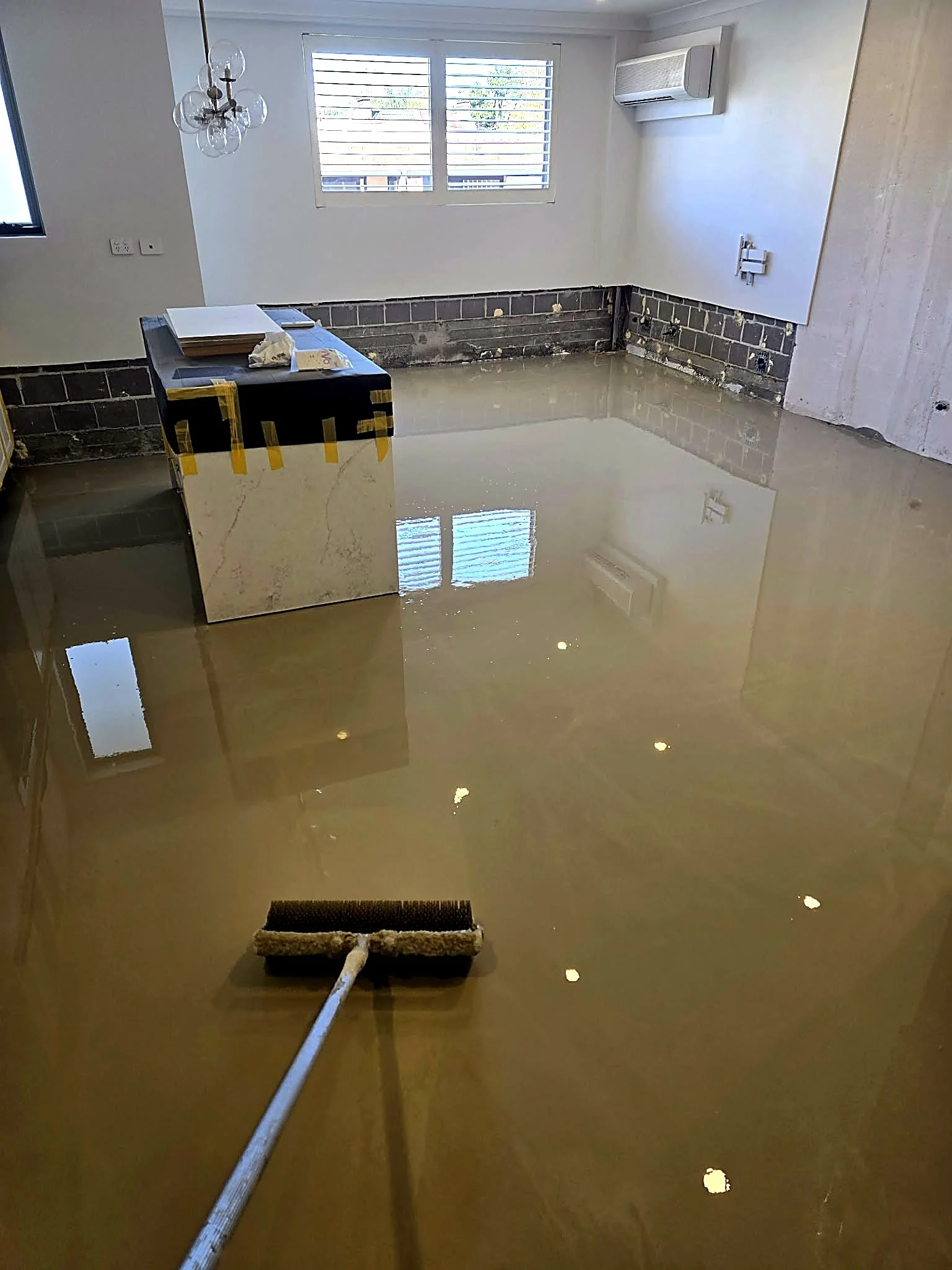 A room under renovation with a partially finished wall, a window with blinds, and a shiny floor. A paint roller with a pole is on the floor, and there are some construction materials on a small table near the wall.