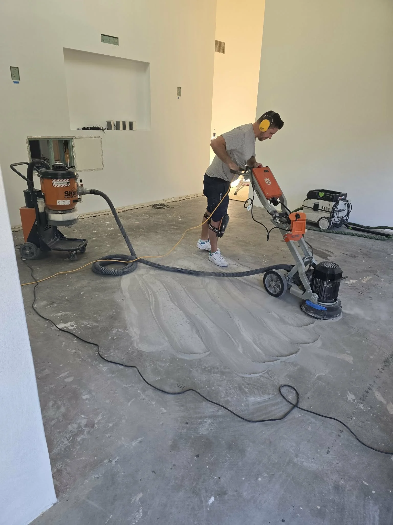 A man wearing gray t-shirt, black shorts, white sneakers, and yellow hearing protection uses floor grinding equipment on concrete floor inside a room with white walls, with various tools and equipment nearby.