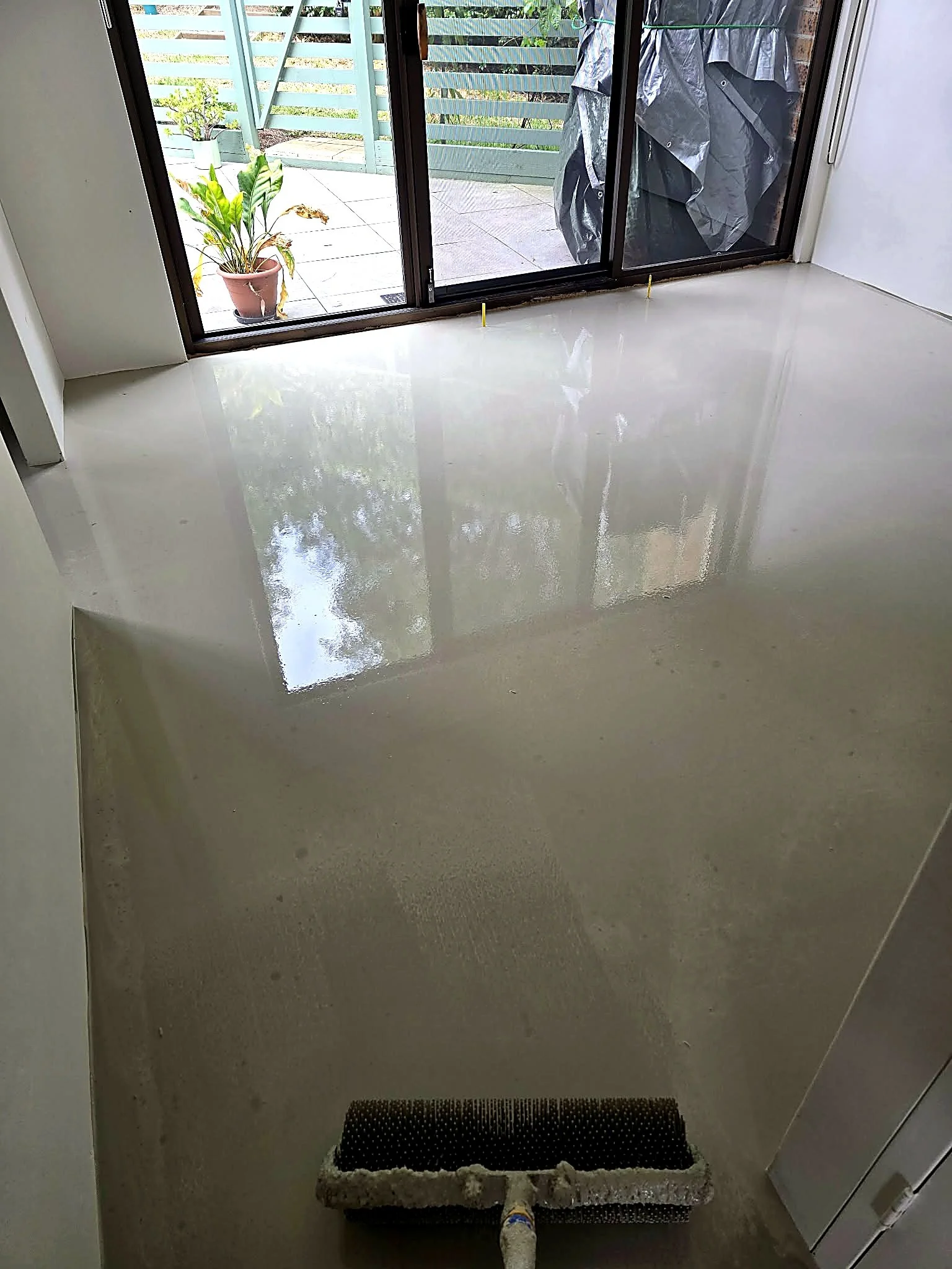 Newly finished shiny floor with an air duct covering in the foreground near a sliding glass door leading to a patio with outdoor plants and drying laundry hanging on a clothesline.