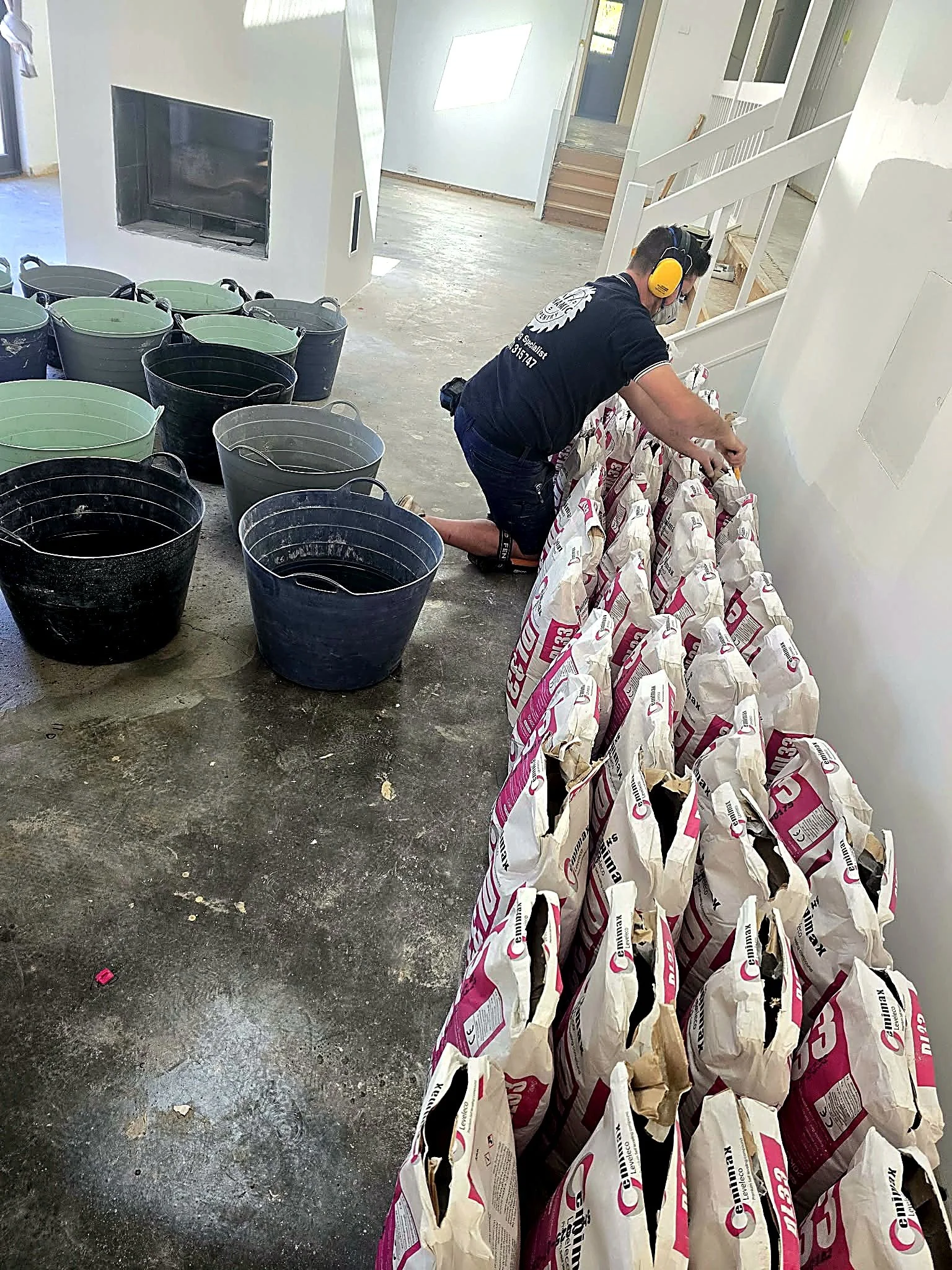A man kneeling on the floor in a room under renovation, wearing yellow ear protection and a mask, working with a row of large bags of cement or similar material, with buckets nearby, in a partially finished interior with white walls and an opening leading to a doorway and staircase.