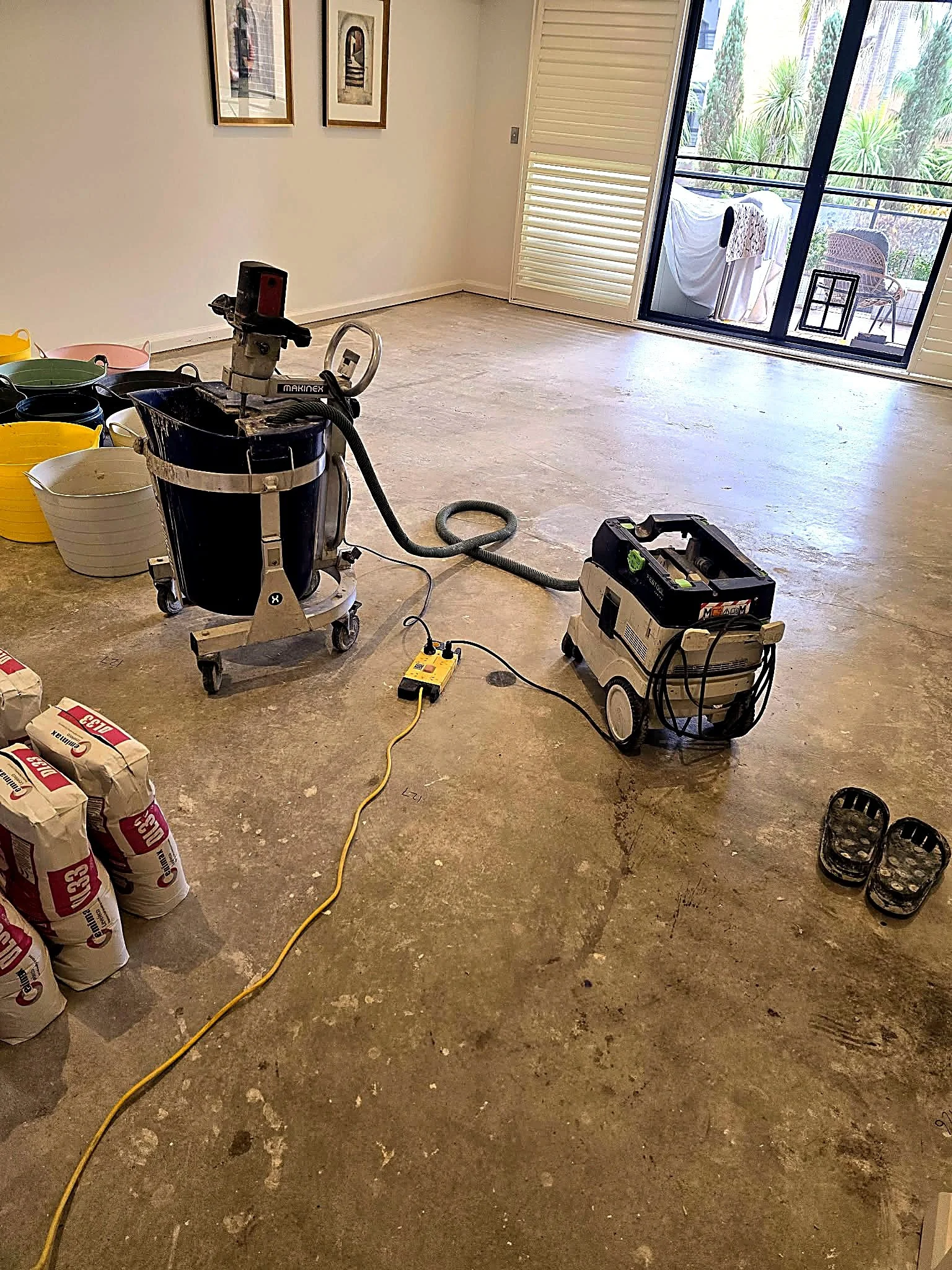 Empty room with a sliding glass door leading to a balcony with outdoor furniture and laundry hanging, construction equipment and materials in the foreground with bags of cement, and slippers on the floor.