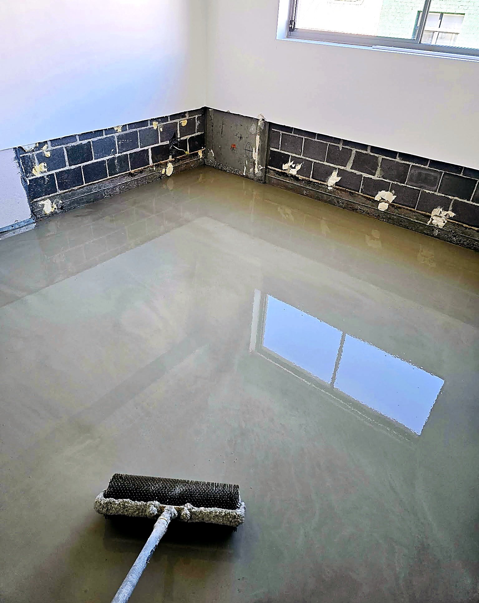 Room with light-colored walls and a large window, showing a wall of a neighboring building outside. The lower part of the walls is exposed brick or concrete with some patches, indicating ongoing construction or renovation. On the floor, a floor sande
