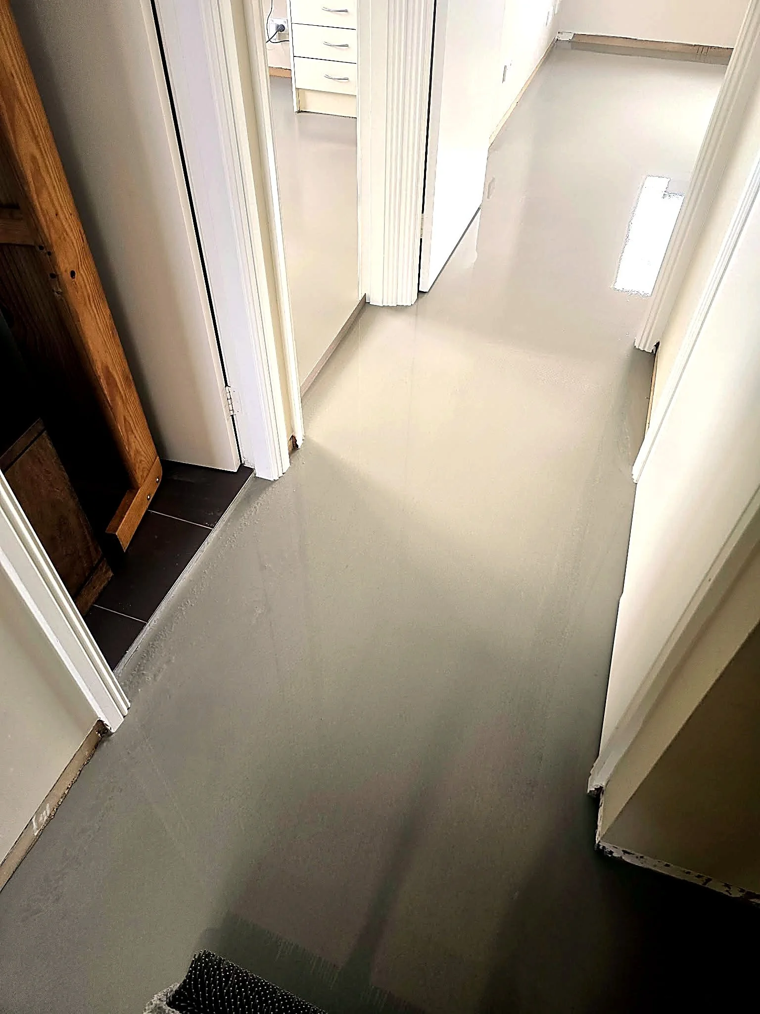 Interior view of a room with a beige wall, an open doorframe, and a gray floor. Part of a wooden piece of furniture and a black woven object are visible in the lower left corner.