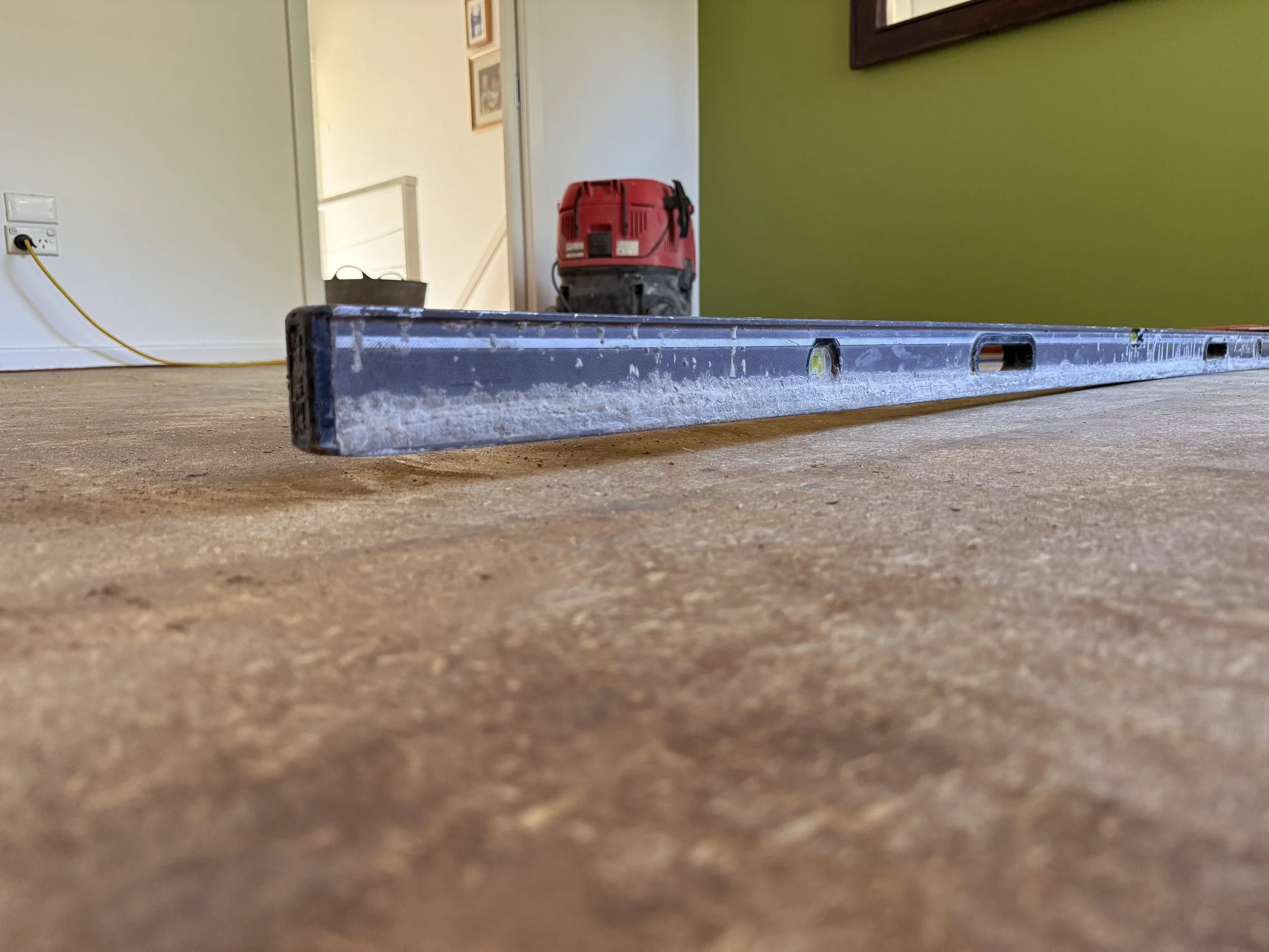A level tool resting on a wooden floor, with a red vacuum cleaner and a green wall in the background.