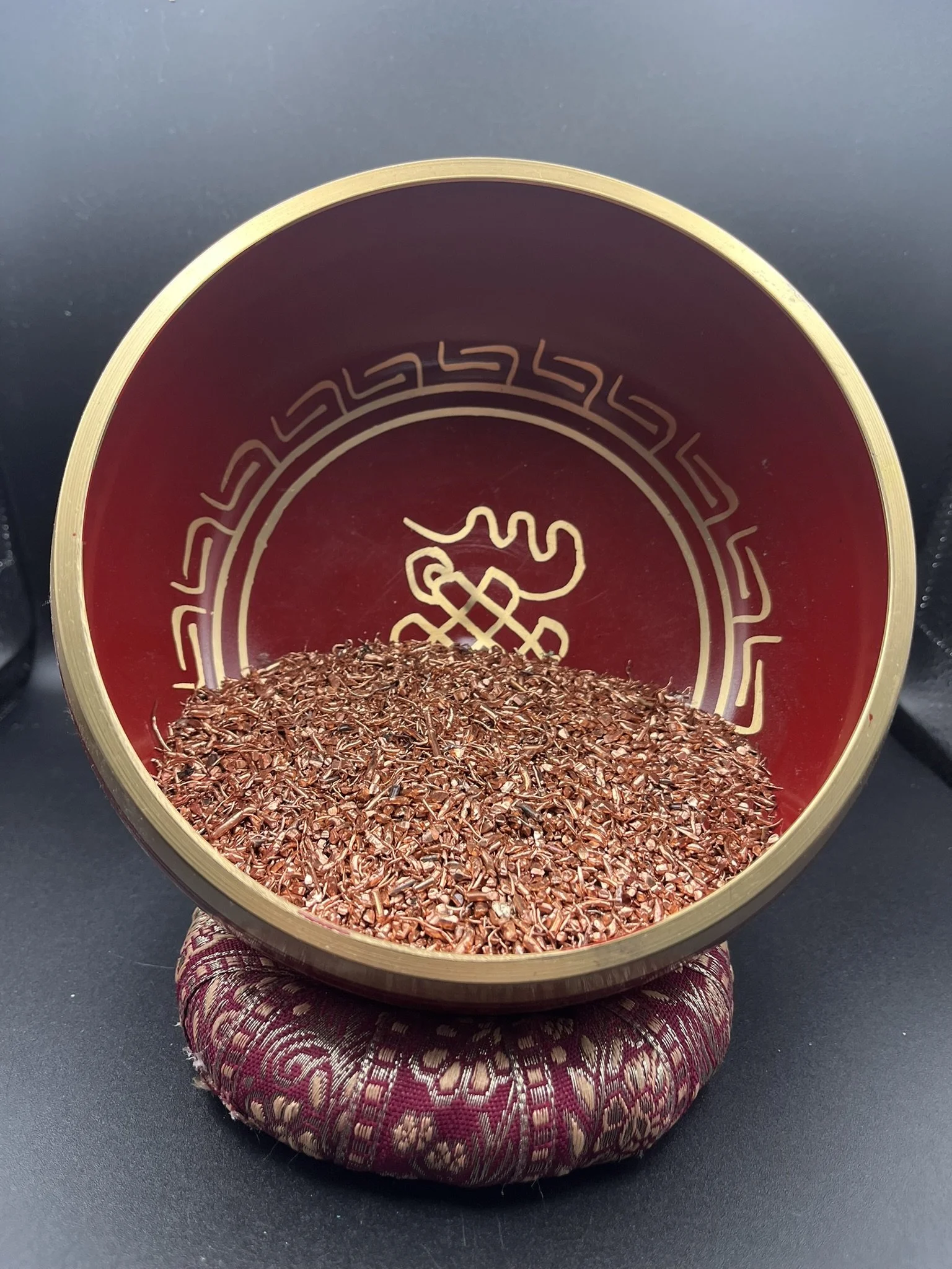 Close-up of a decorative container filled with dried red chili flakes, placed on a patterned fabric cushion on a dark surface.