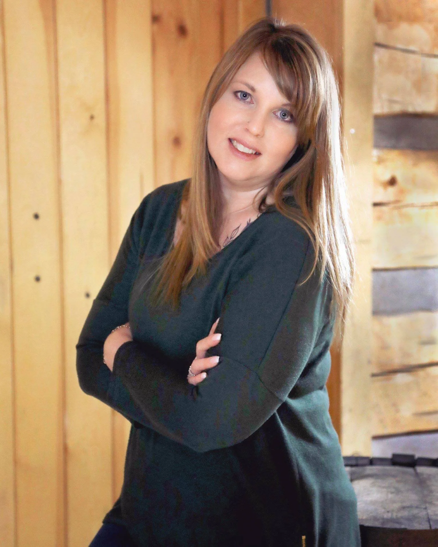 A woman with long brown hair, wearing a black top, posing with her arms crossed in front of a wooden wall.