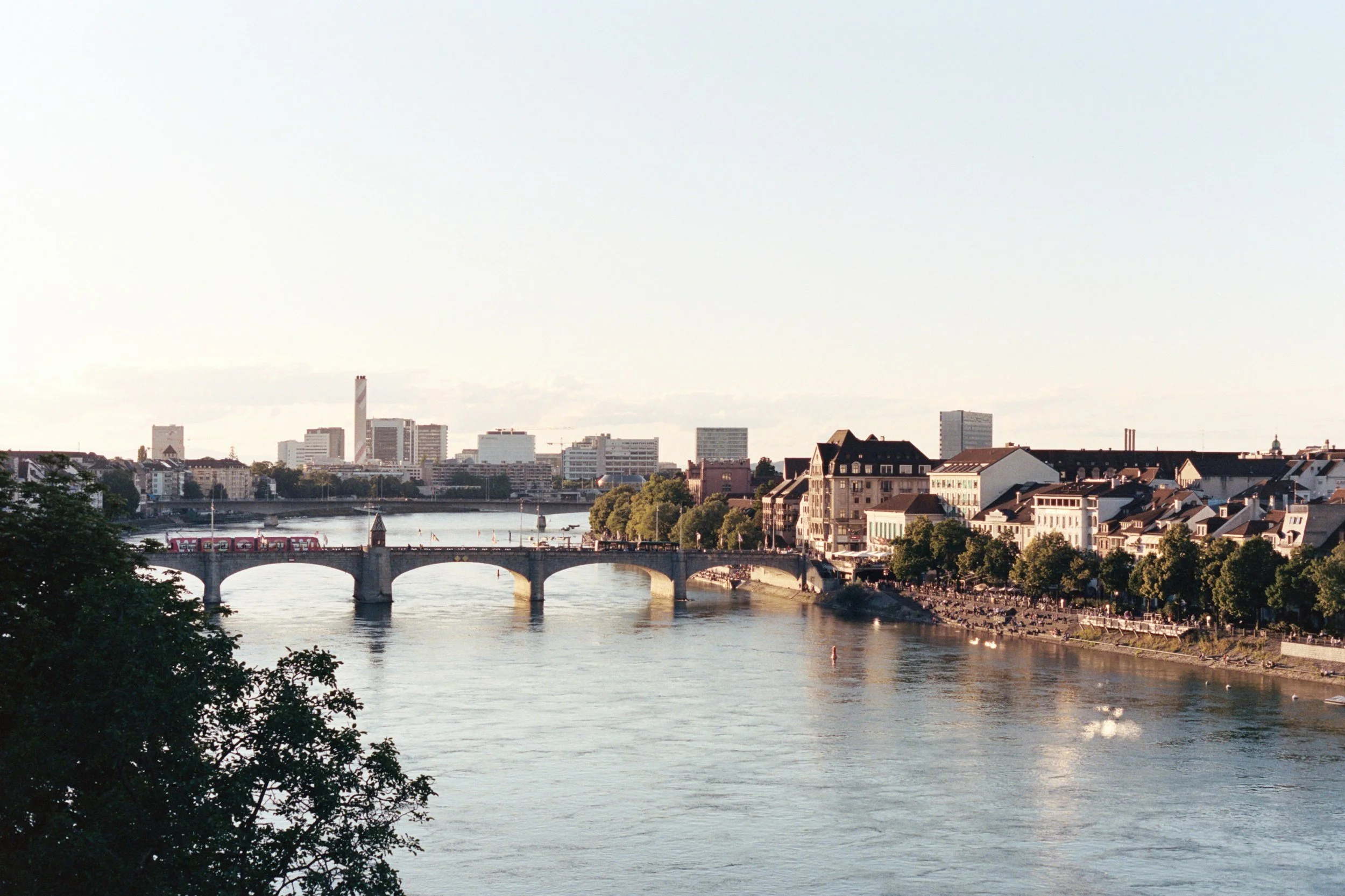 A cityscape featuring a river with a bridge, modern buildings in the background, and a historic building with a pointed roof in the foreground.