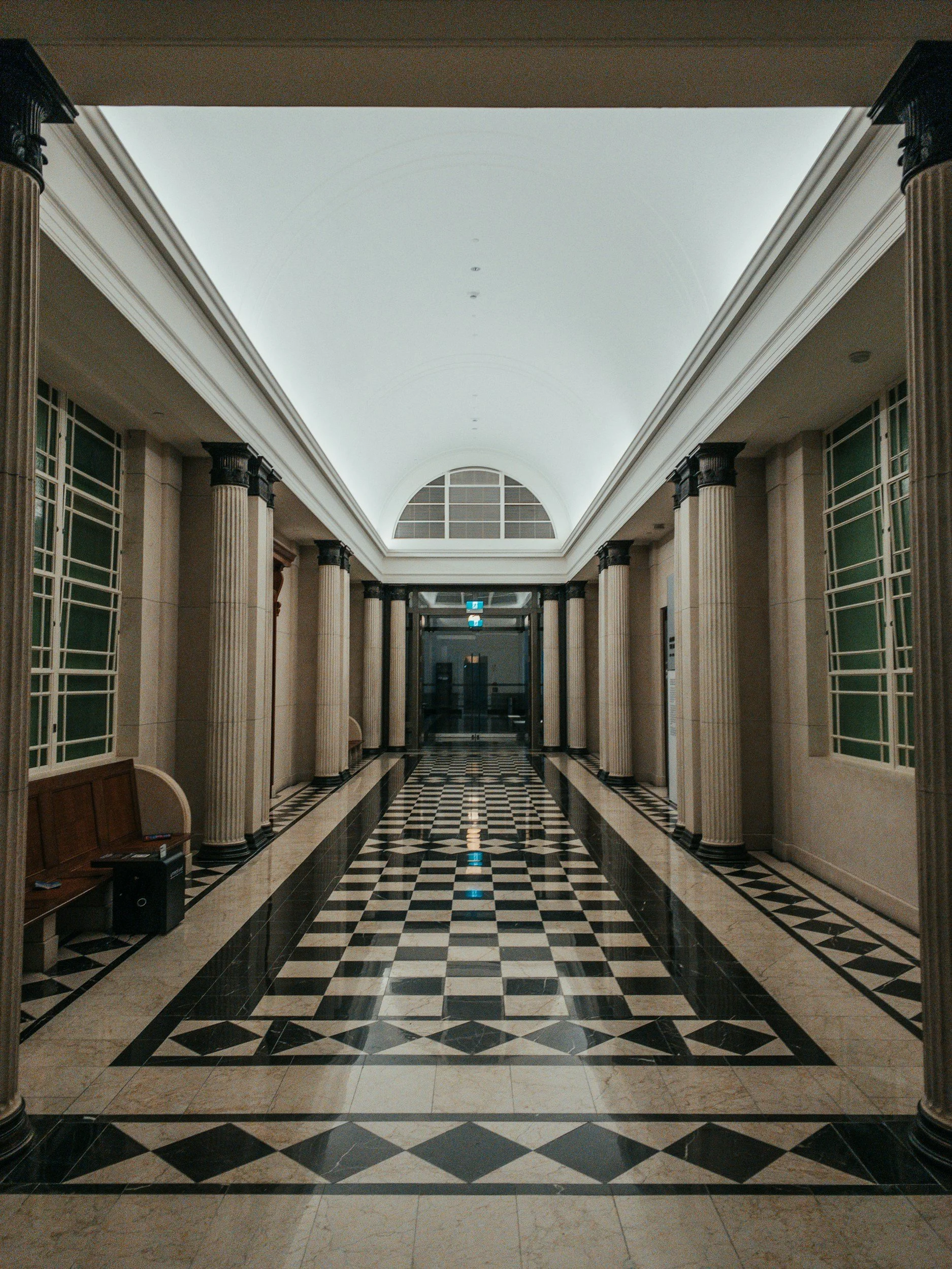 An elegant, symmetrical hallway with a checkered black and beige marble floor, tall beige columns with black capitals, and large windows with green panes on both sides. The ceiling is white with a vaulted design and a large arched window at the end.