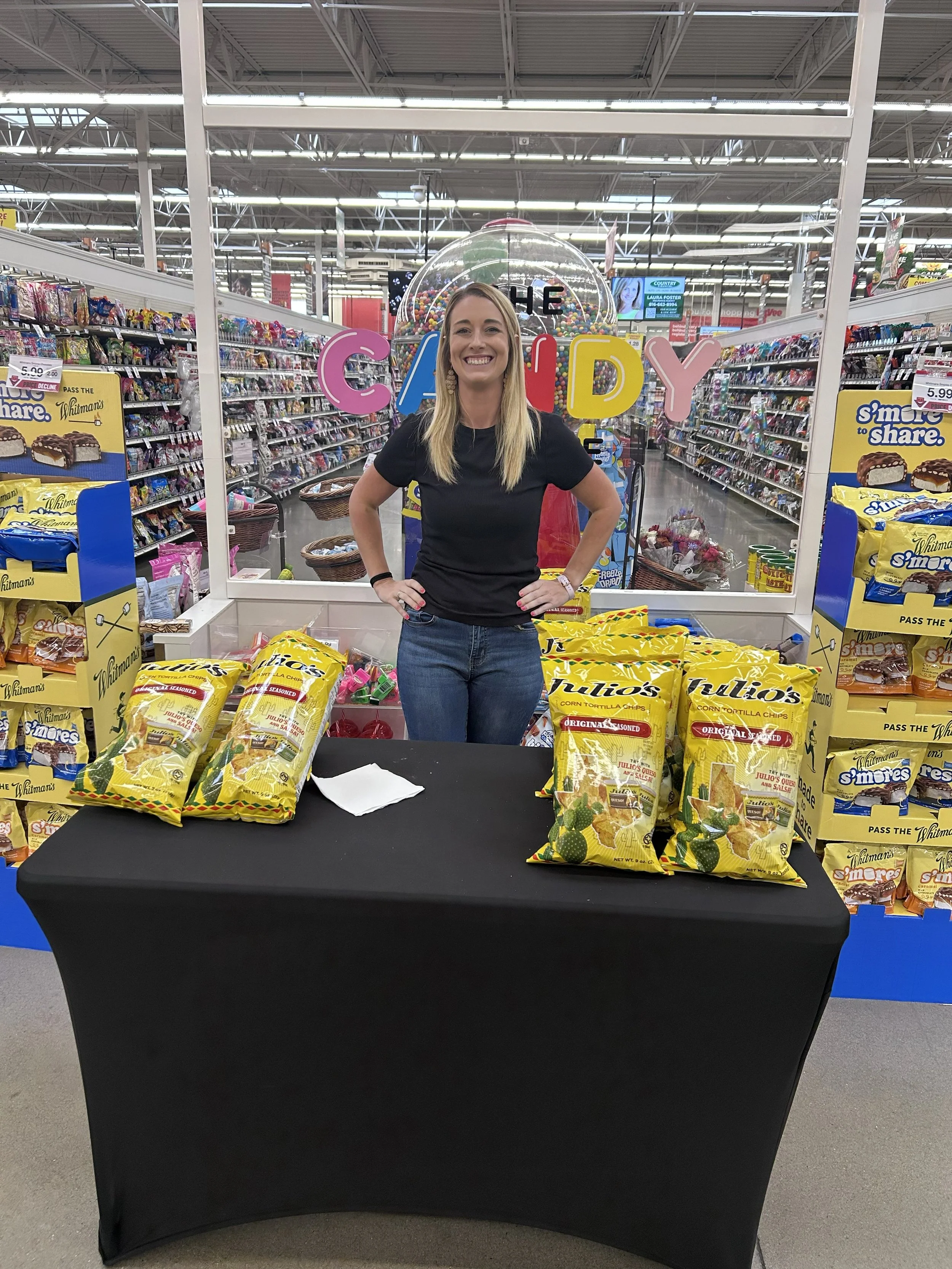 A woman standing behind a display table of bags of Tortilla Chips inside a grocery store, with shelves of various snack products and candy in the background.